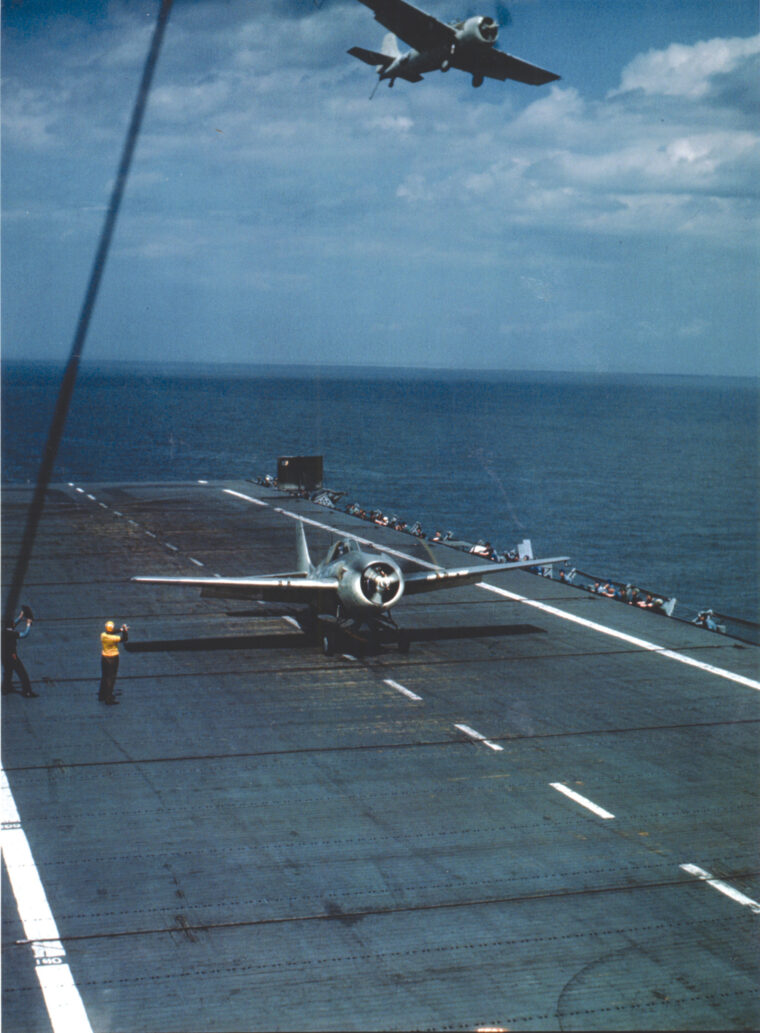 Navy personnel look on from the edge of a carrier deck as an F4F-3A prepares for takeoff.