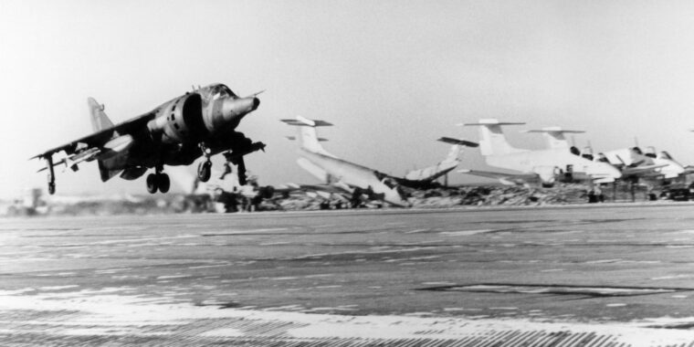 A Sea Harrier takes off from the airfield at Port Stanley during Falklands war. In the background is a destroyed Argentinian Air Force IA 58 Pucará counter-insurgency (COIN) aircraft.