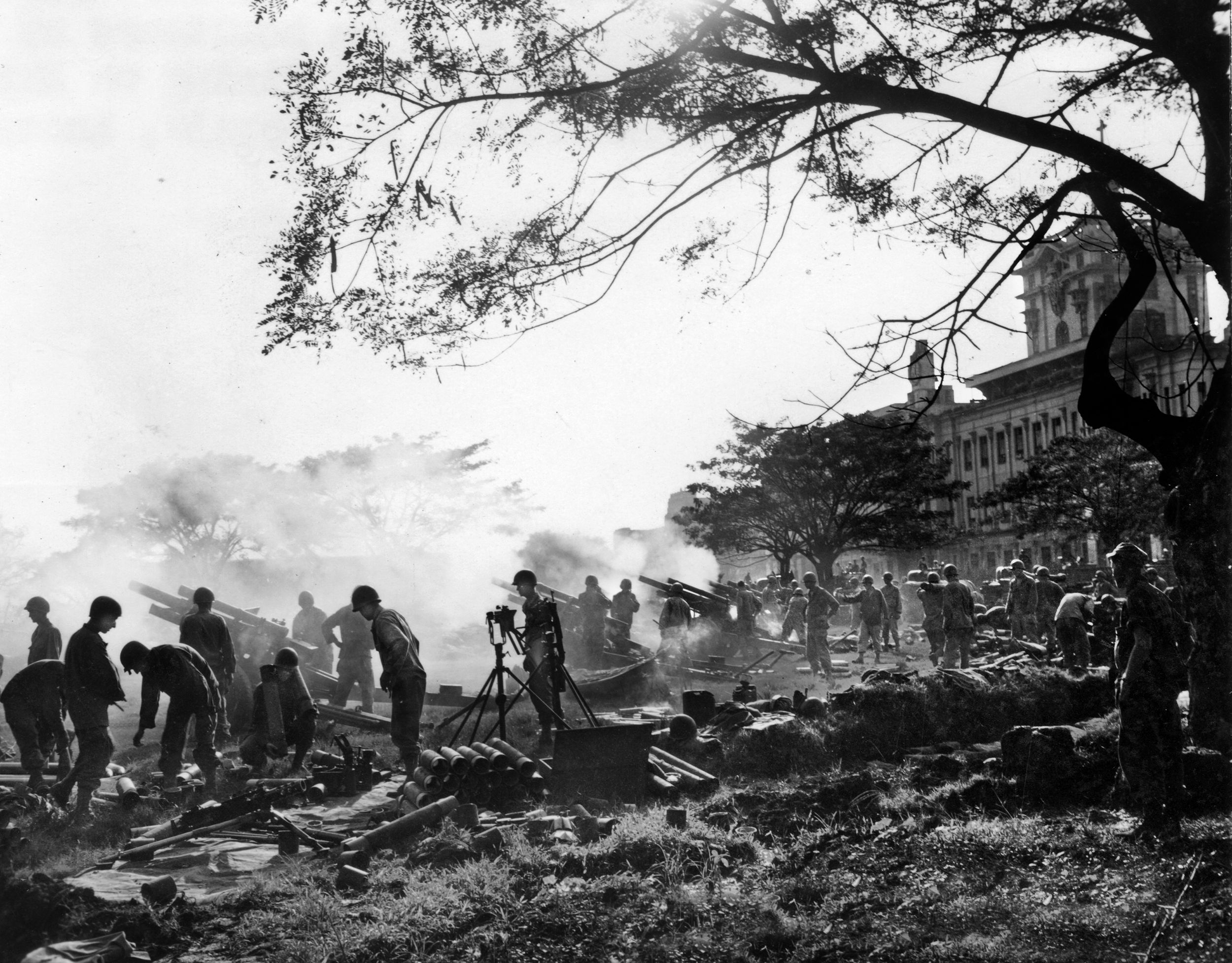 On February 5, 1945, American soldiers service their 105mm howitzers, firing against Japanese positions in the vicinity of Manila, from the grounds of Santo Tomas, where a university had been turned into a detention center for prisoners.