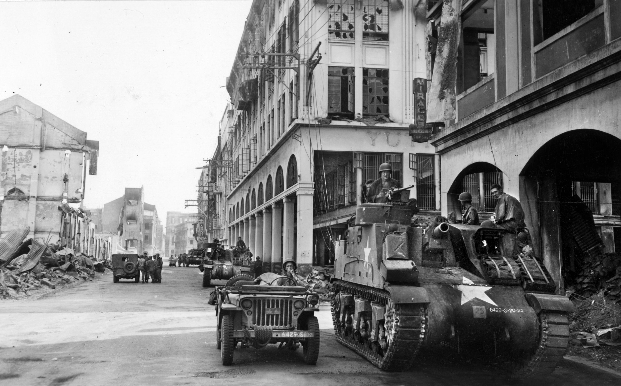 American soldiers pause in a Manila street prior to advancing toward Japanese positions. A self-propelled 105mm howitzer is seen in this photo of February 17, 1945, along with a jeep, while a string of  other vehicles extends into the distance.