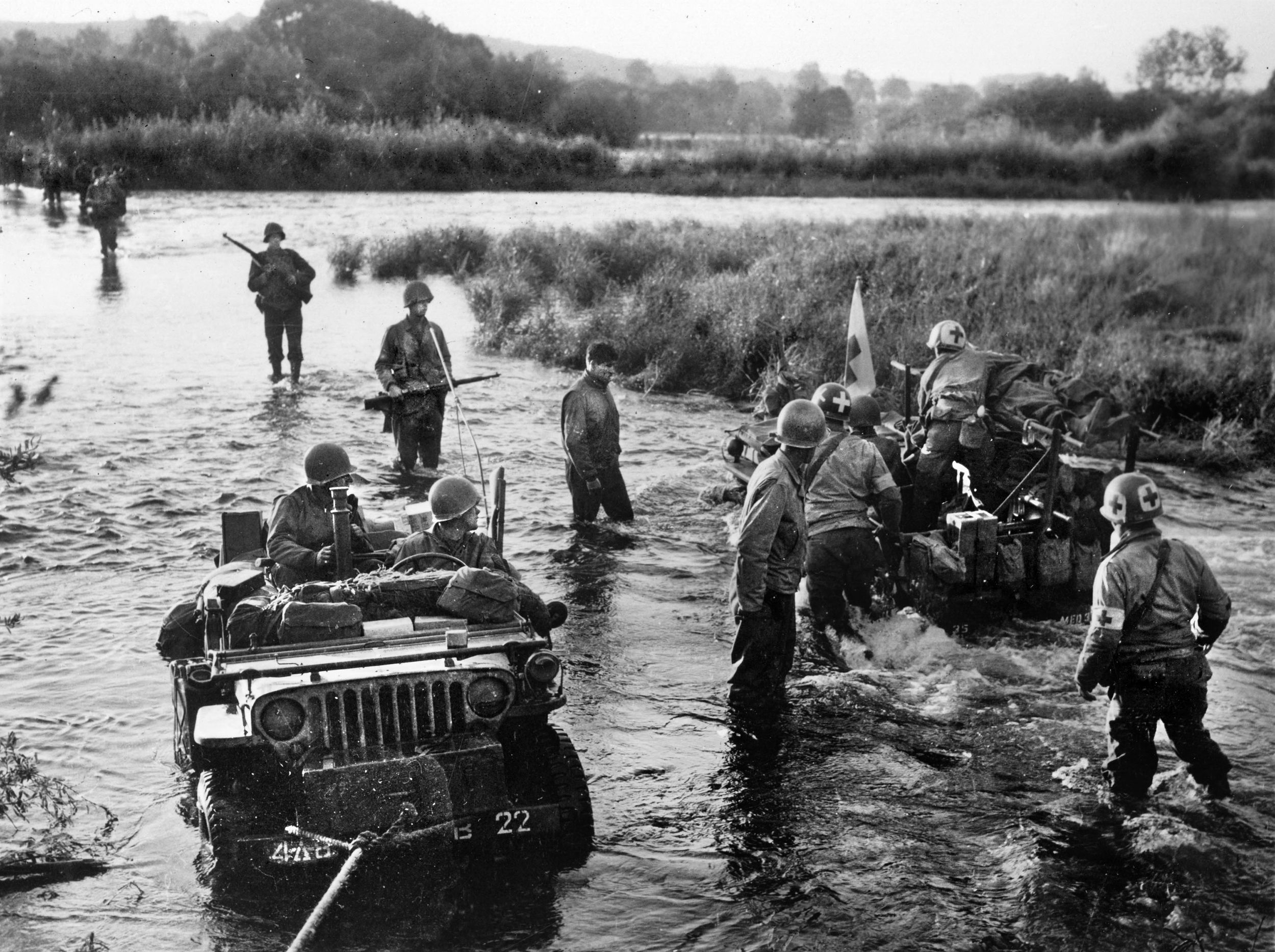 American soldiers wade and drive a Jeep across the Moselle River in September 1944, pausing to watch another vehicle move in the opposite direction, probably carrying wounded. Note the Red Cross identification and the presence of medics.