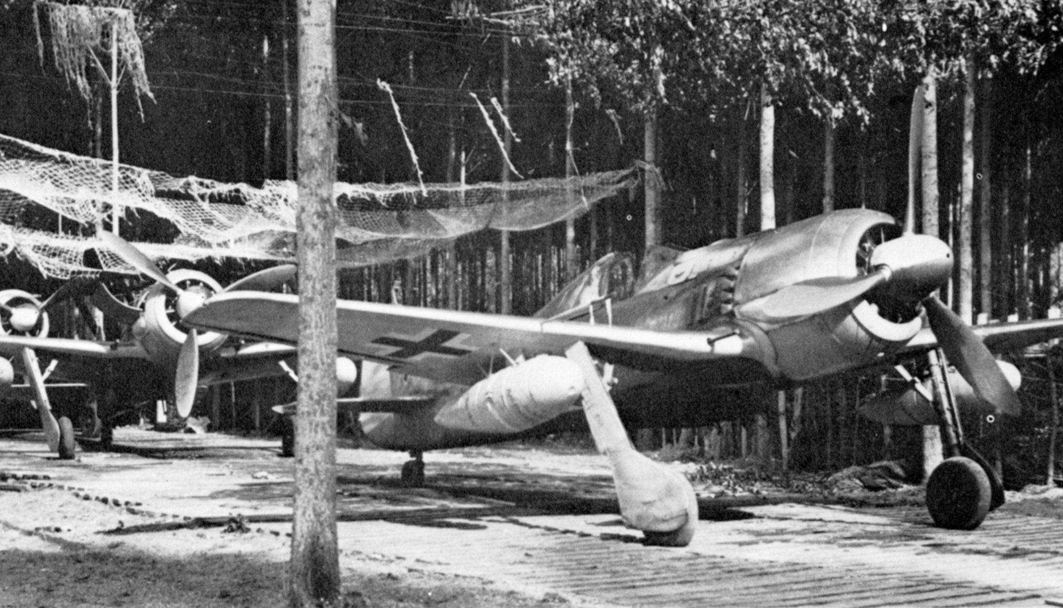 Hoping to avoid the eyes of Allied fighter-bombers, German fighters sit under the cover of trees and camouflage netting. 
