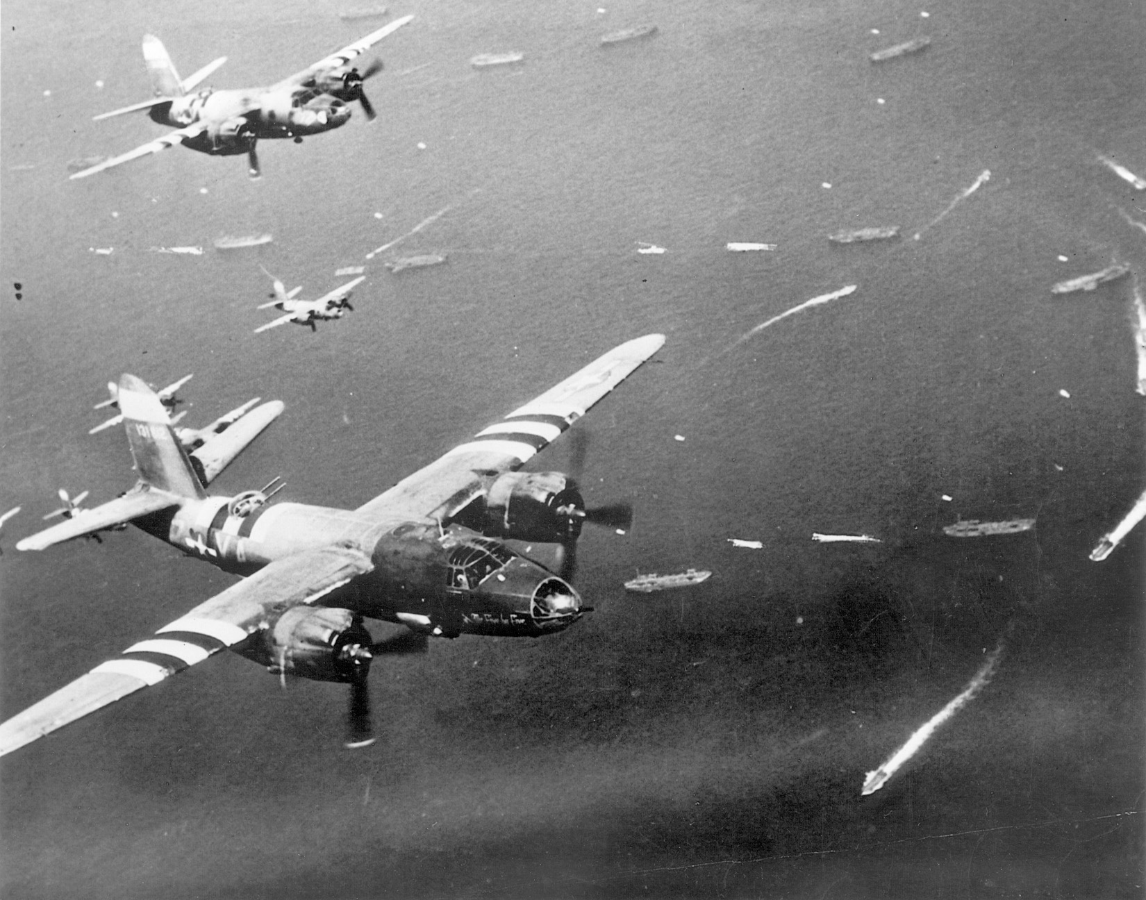 Painted with black and white recognition stripes for the D-day invasion, twin-engined Martin B-26 bombers head toward targets in France. Ships of the Allied invasion force mill about in the English Channel below.