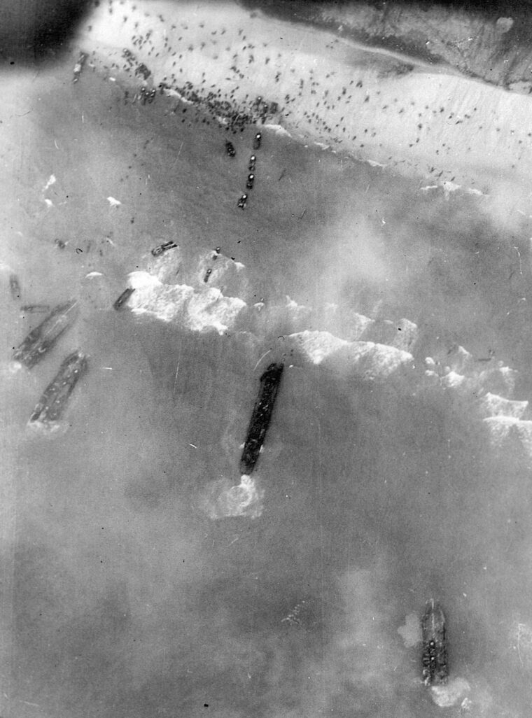 In this aerial view of the initial D-day landings on the Normandy coast, American troops can be seen struggling toward shore from their landing craft amid a storm of German small arms fire. Some U.S. casualties lie wounded or dead in the surf and at the water’s edge. 
