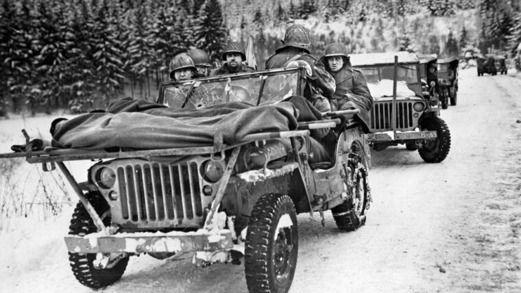 U.S. Army medical personnel use stretchers across the hood of a Jeep to evacuate wounded soldiers from the front line in Bastogne, Belgium, during heavy action—evidenced by the bullet hole in the Jeep’s windshield.