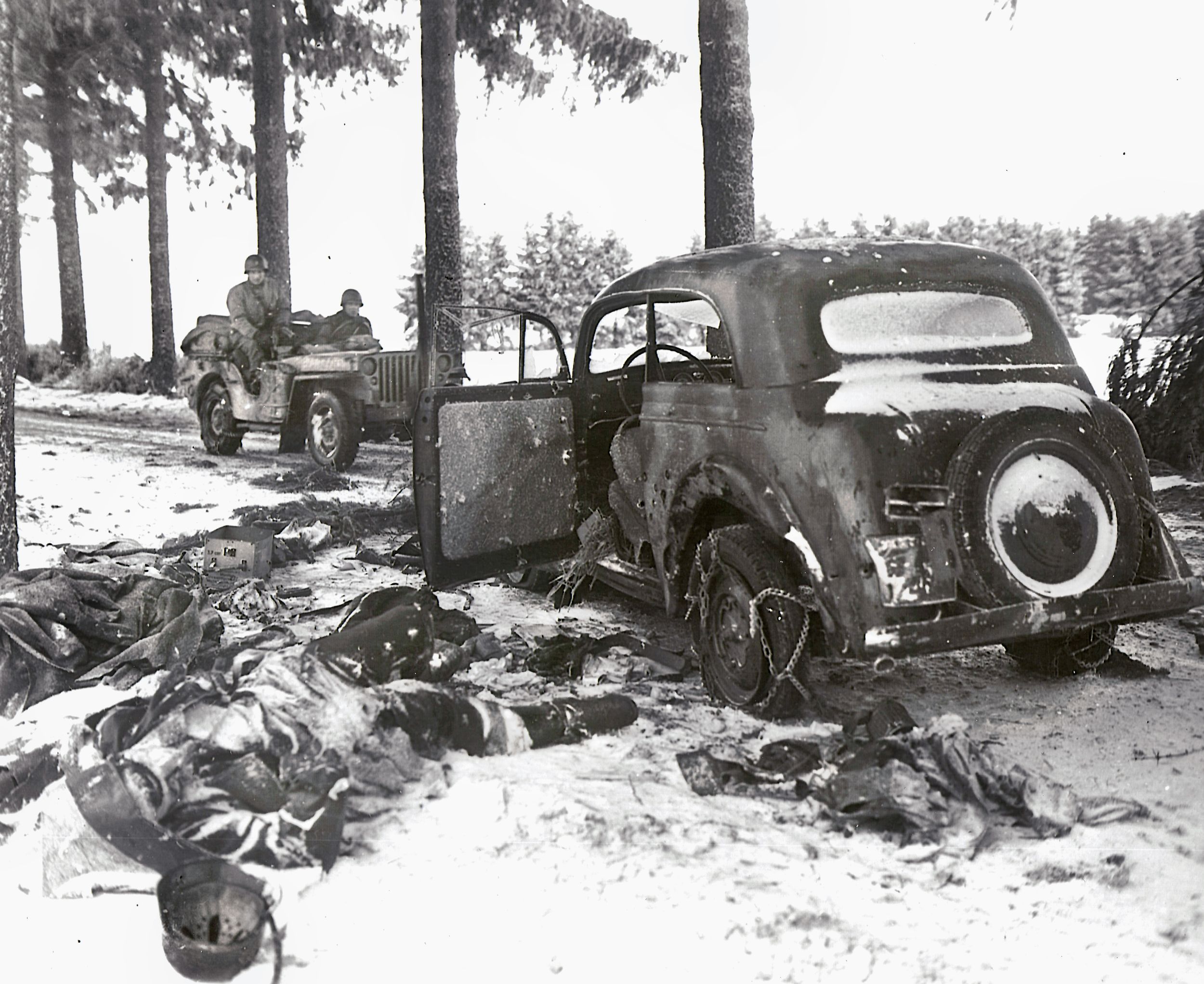 American soldiers drive by a destroyed German vehicle and the bodies of its former occupants during the Battle of the Bulge. U.S. Army PFC Frank Cohn and his team were suspected of being German spies when they were stopped and interrogated at an American checkpoint in Belgium.