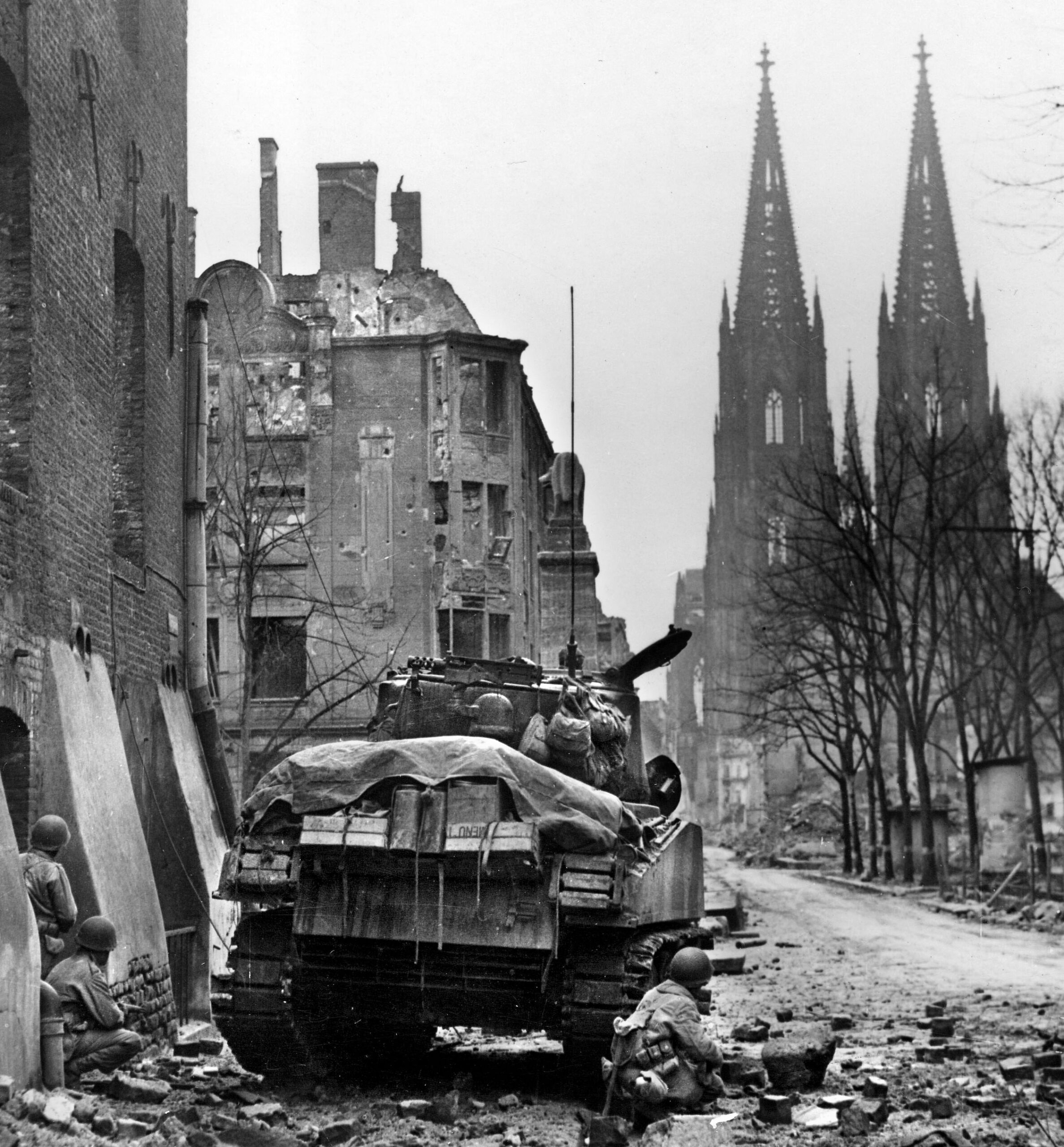 U.S. soldiers squat behind a tank and building as they fight to capture Cologne on March 6, 1945. PFC Cohn almost lost his life to a German mortar barrage in the city.