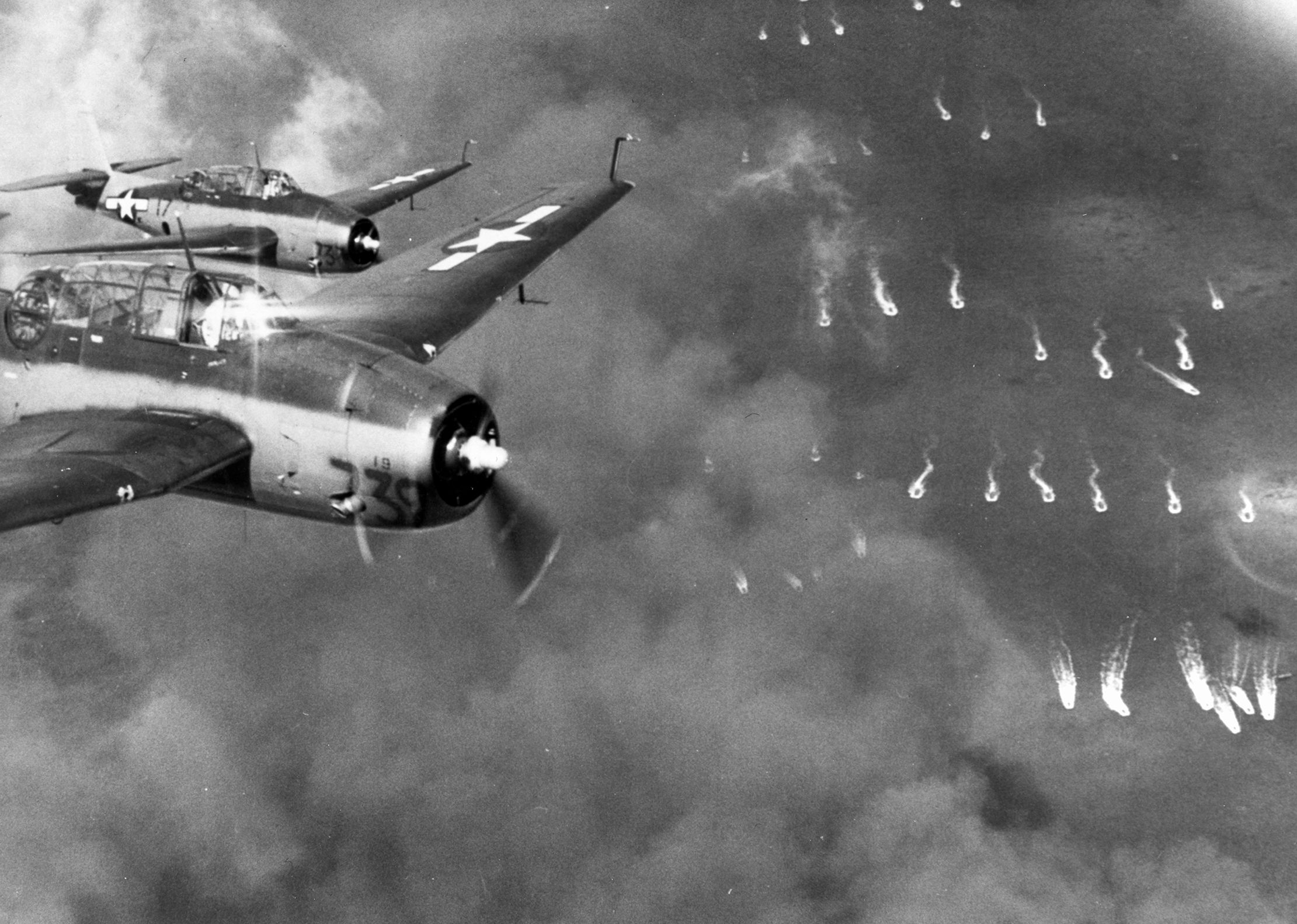 A pair of Grumman TBF Avenger bombers from the escort carrier USS Coral Sea fly near Butaritari during Operation Galvanic, November 20, 1943. The wakes of landing craft turning toward shore and the fires burning from preinvasion bombardment are visible below.