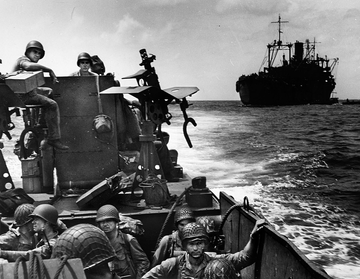 A tank lighter manned by U.S. Coast Guard personnel makes it way toward shore at Makin while hauling a heavy gun and tractor. The ship in the background is a combat transport also manned by Coast Guardsmen in service with the U.S. Navy task force. As this photo was taken, American bombers were hitting targets on the islet of Butaritari. 