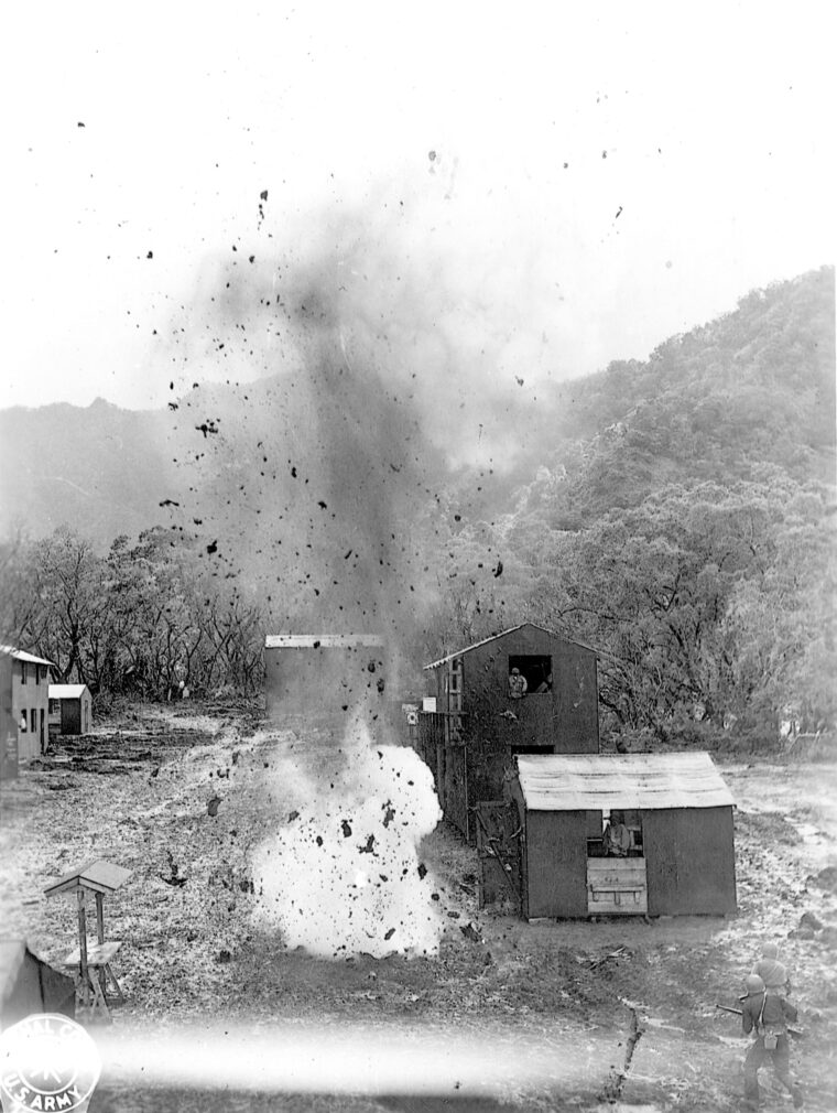 A bomb detonates in the street of a mockup Japanese village as men work their way through it during an exercise in house-to-house fighting. Note the dummies in the windows of the houses on the right.