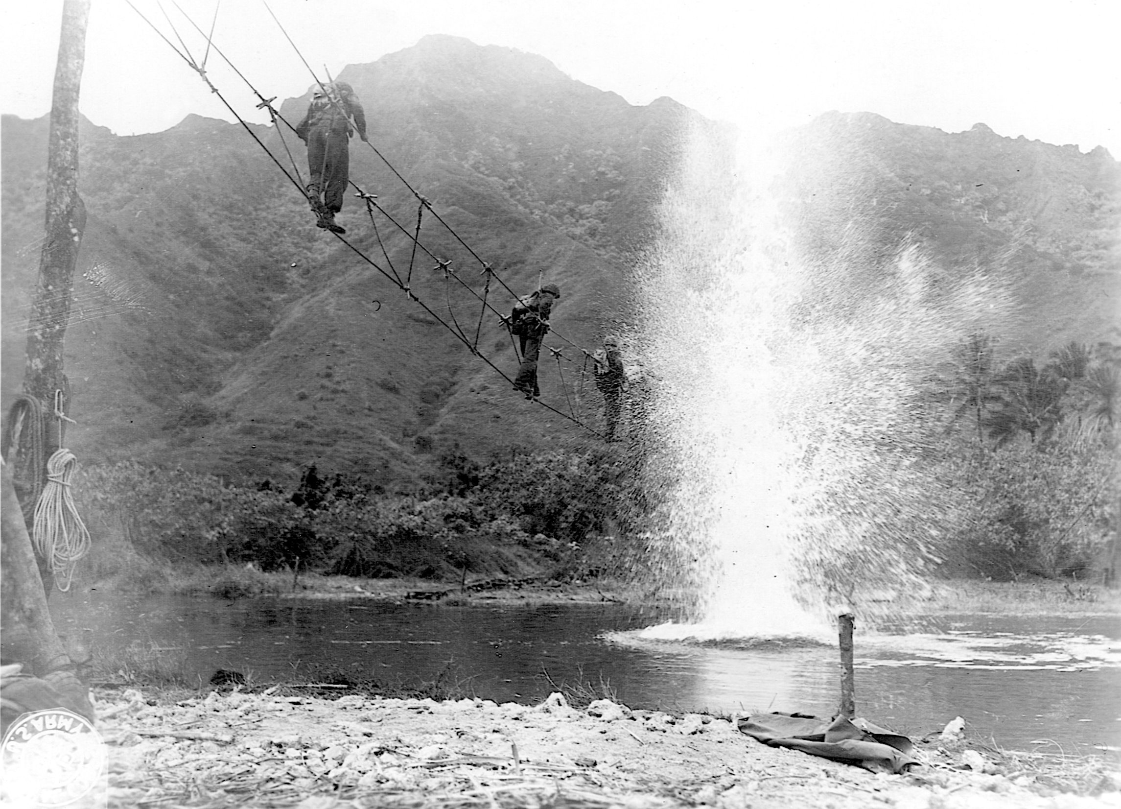The splashes of exploding dynamite greet soldiers crossing a stream on a narrow toggle bridge.