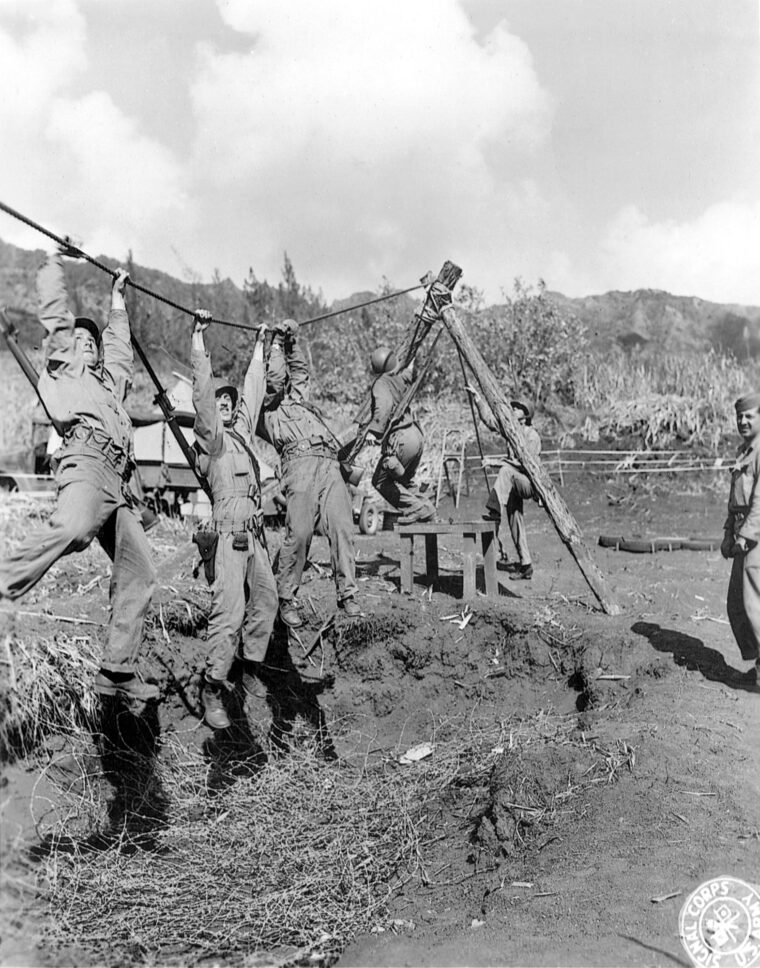 Hand over hand, soldiers cross a trench filled with barbed wire. The men endured strenuous tests, some under fire, in preparation for combat in the Pacific.