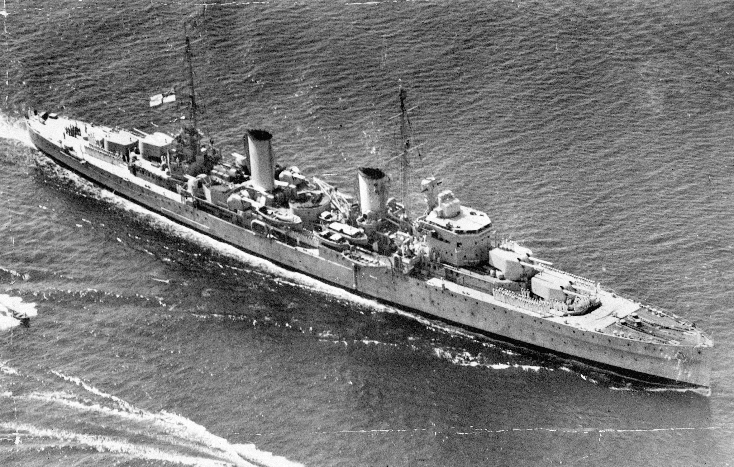 In a 1940 aerial view, HMAS Perth is shown underway. The fire director for the main armament may be seen behind the bridge, while the high angle control position for the 4-inch main batteries is above.  Searchlights flank the fore funnel. The eight 4-inch Mark XVI guns are grouped in twin Mark XIX mounts. 