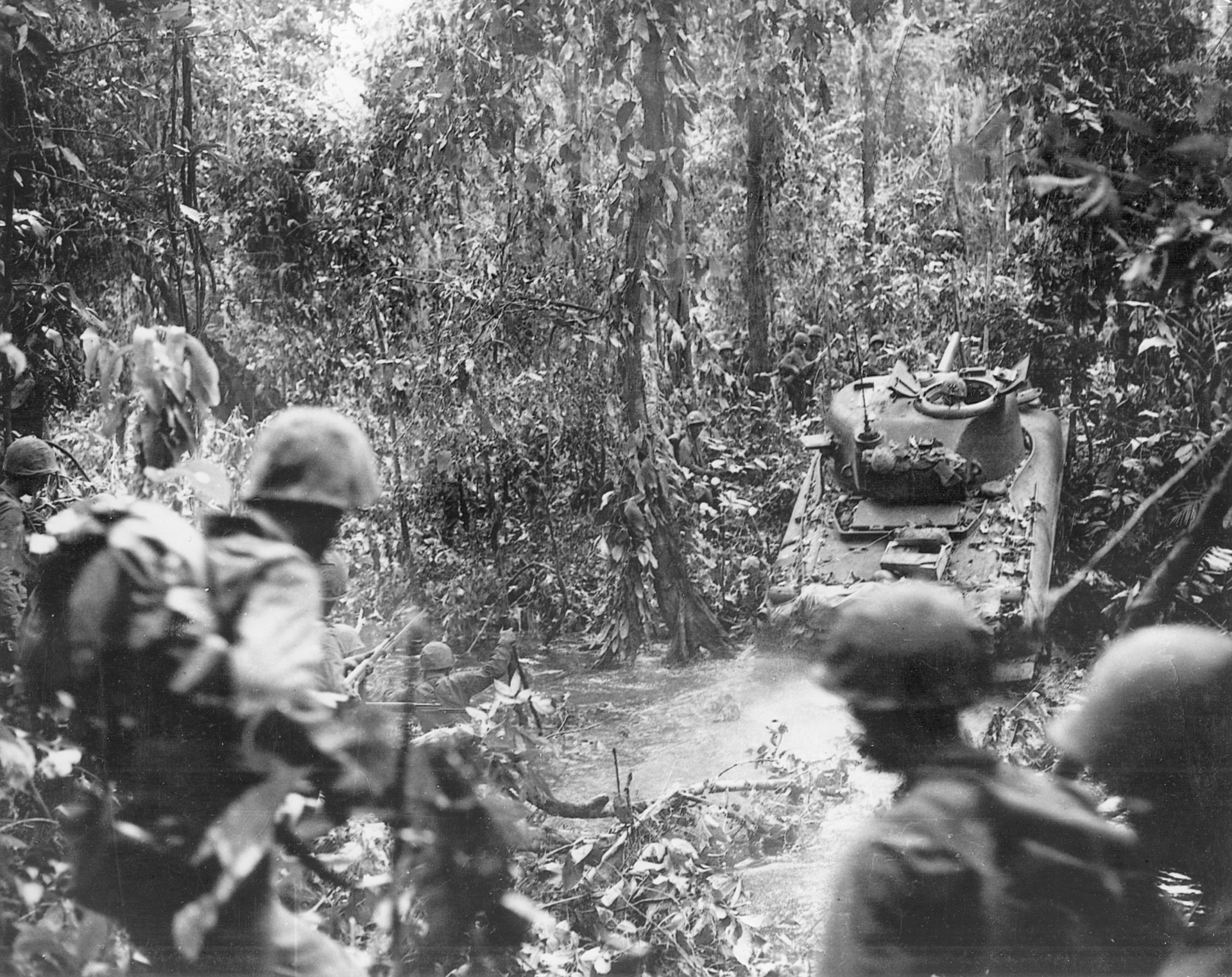 A Sherman tank leads Marines across a stream and up an embankment in the steamy New Guinea jungle.