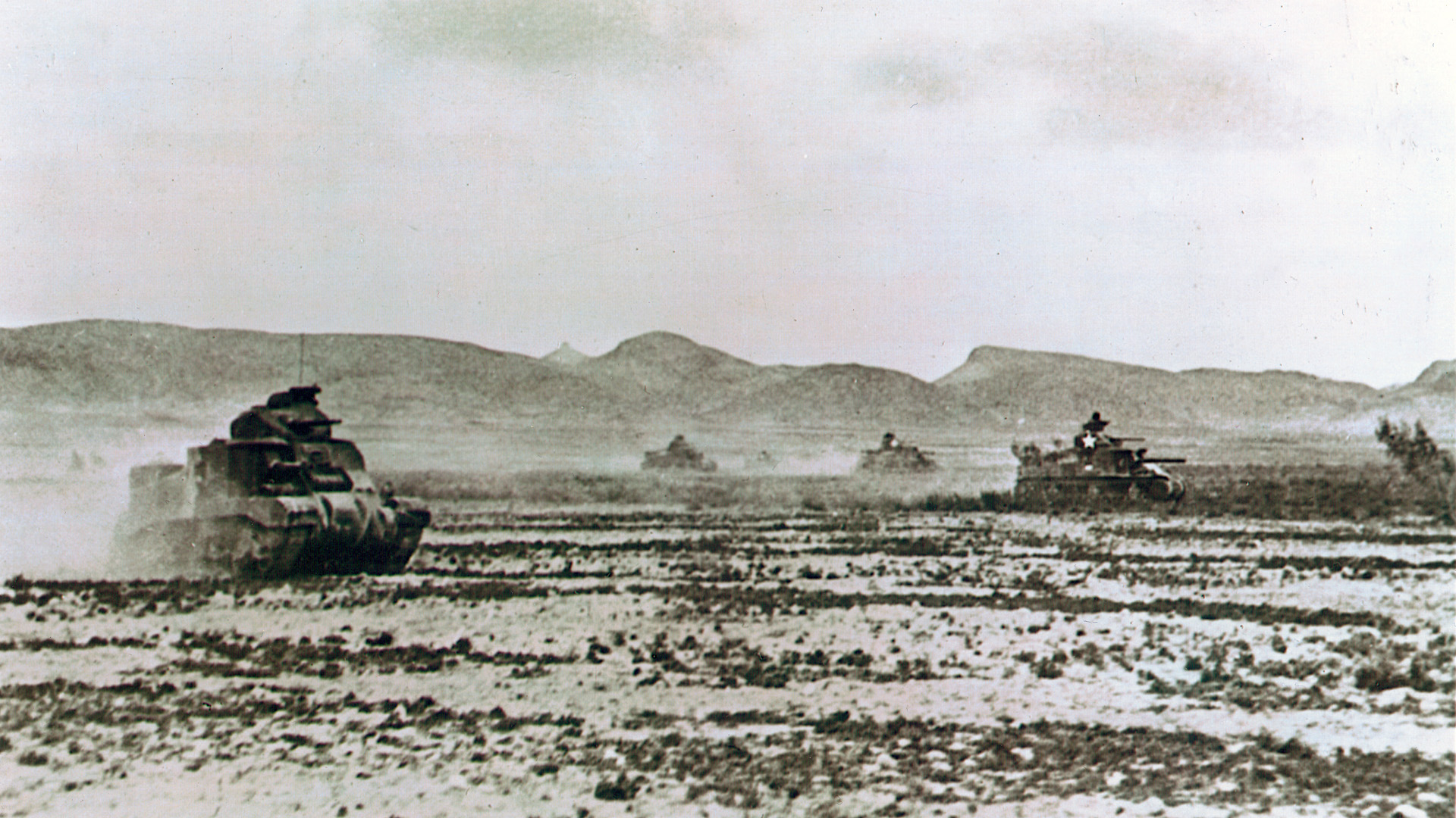 American M3 Lee tanks, with hull-mounted 75mm canon and 37mm guns in turrets above, raise clouds of dust as they race across the Tunisian desert.