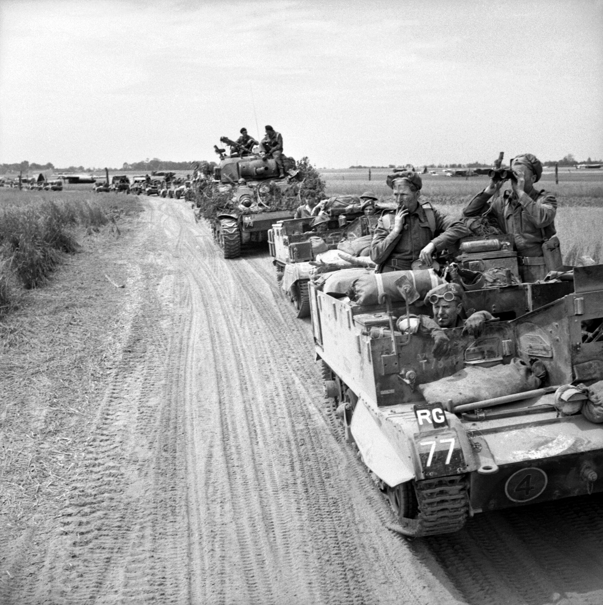 During the opening day of Operation Goodwood, British troops aboard Bren gun carriers advance toward their objective near Caen. A Sherman tank being utilized as an artillery unit headquarters accompanies the infantrymen.