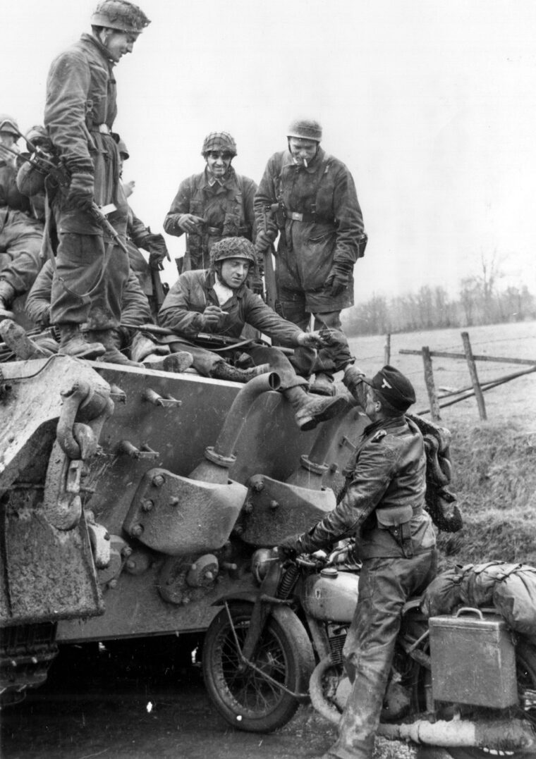 A German Fallschirmjäger hands a cigarette to a messenger who has just come up on a motorcycle during the advance of the 501st Heavy SS Battalion in the Battle of the Bulge. The German paratroopers pictured aboard the Tiger tank were  attached to Kampfgruppe Peiper. The German battle group led by SS Lt. Col. Joachim Peiper was intent on capturing bridges at Trois-Ponts, Belgium, but heroic American combat engineers thwarted his plan.