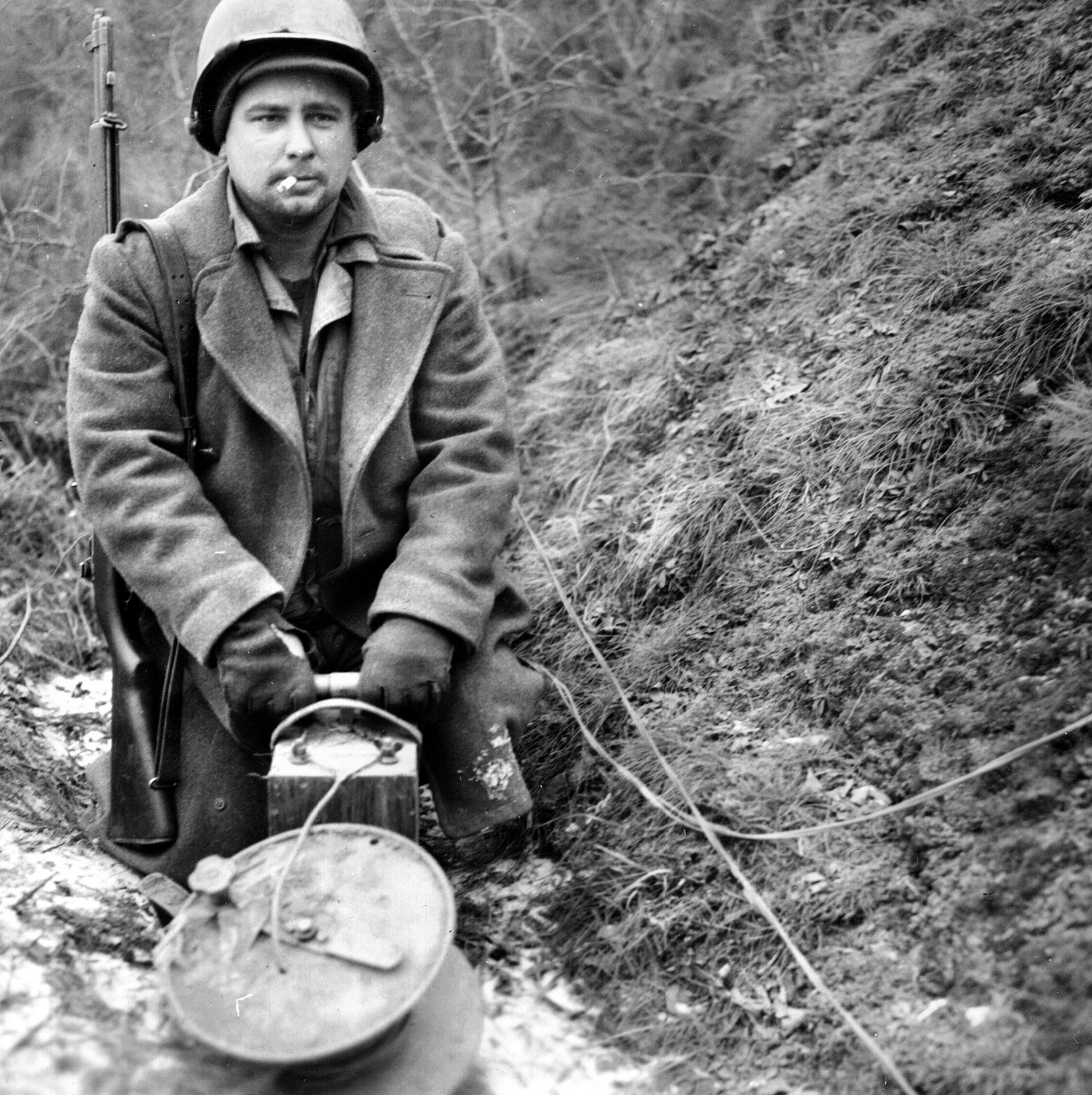 A U.S. Army combat engineer stands with hands on the plunger of a detonator box. Engineers have just completed wiring explosives to large trees along a roadway, expecting the detonation to fell the trees and block the progress of the Nazi spearheads toward the Meuse River during the Battle of the Bulge.