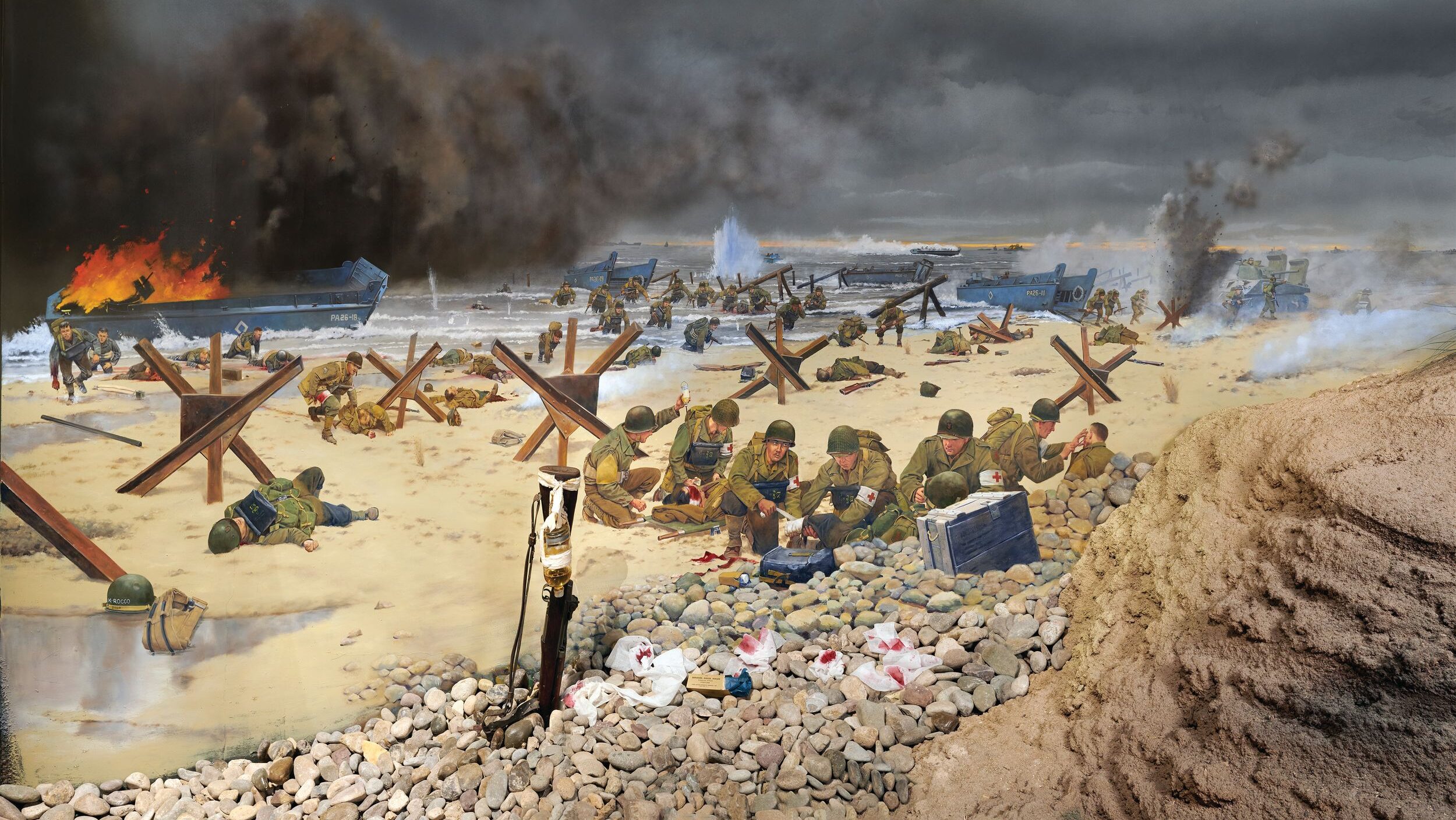 Medics, who have lost most of their supplies, still treat the wounded on Omaha Beach on June 6, 1944, in this mural by Keith Rocco. Center, left, bandaging a soldier’s leg is medic Charles Norman Shay, of the Penobscot Indian Nation, who received a Silver Star. Further to the left is wounded African American medic Waverly Woodson, Jr., who is helping a fellow soldier crawl forward. Woodson, who treated more than 200 men before collapsing after 30 hours, received a Bronze Star.