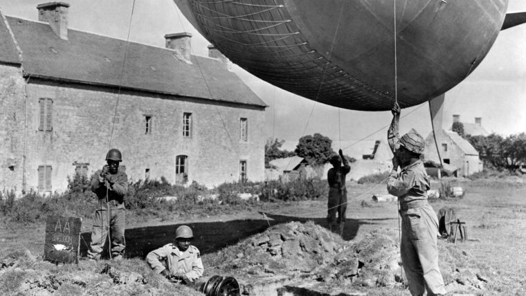 Soldiers of the 320th Barrage Balloon Battalion work on the moorings of a barrage balloon on the coast of Normandy after D-Day. The balloons were made of two-ply cotton fabric impregnated with vulcanized or synthetic rubber, then coated with aluminum. Typically, the balloons were raised in the evening after Allied aircraft had returned to bases in England.