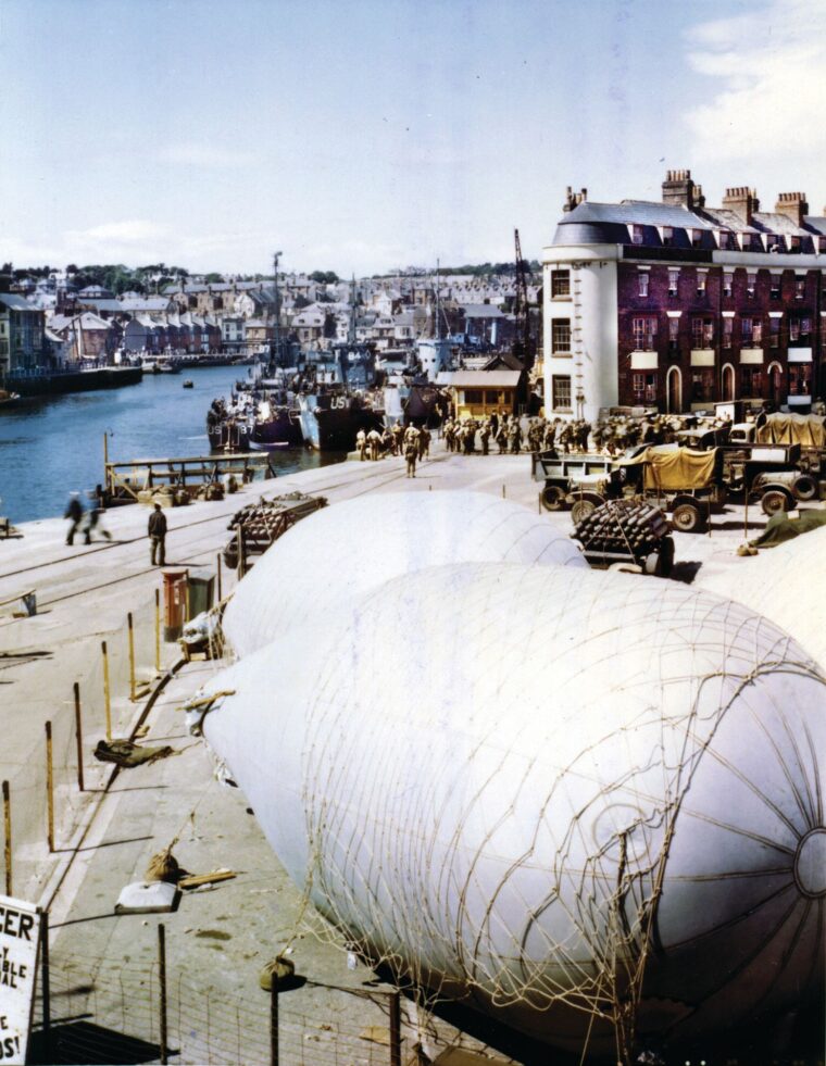 While Allied infantrymen board transport craft that will take them to the invasion beaches on D-Day, a barrage balloon is prepared for tethering to a ship that will participate in the landings. The 320th Barrage Balloon Battalion, an African American unit, served with distinction on D-Day and during the Normandy campaign. 