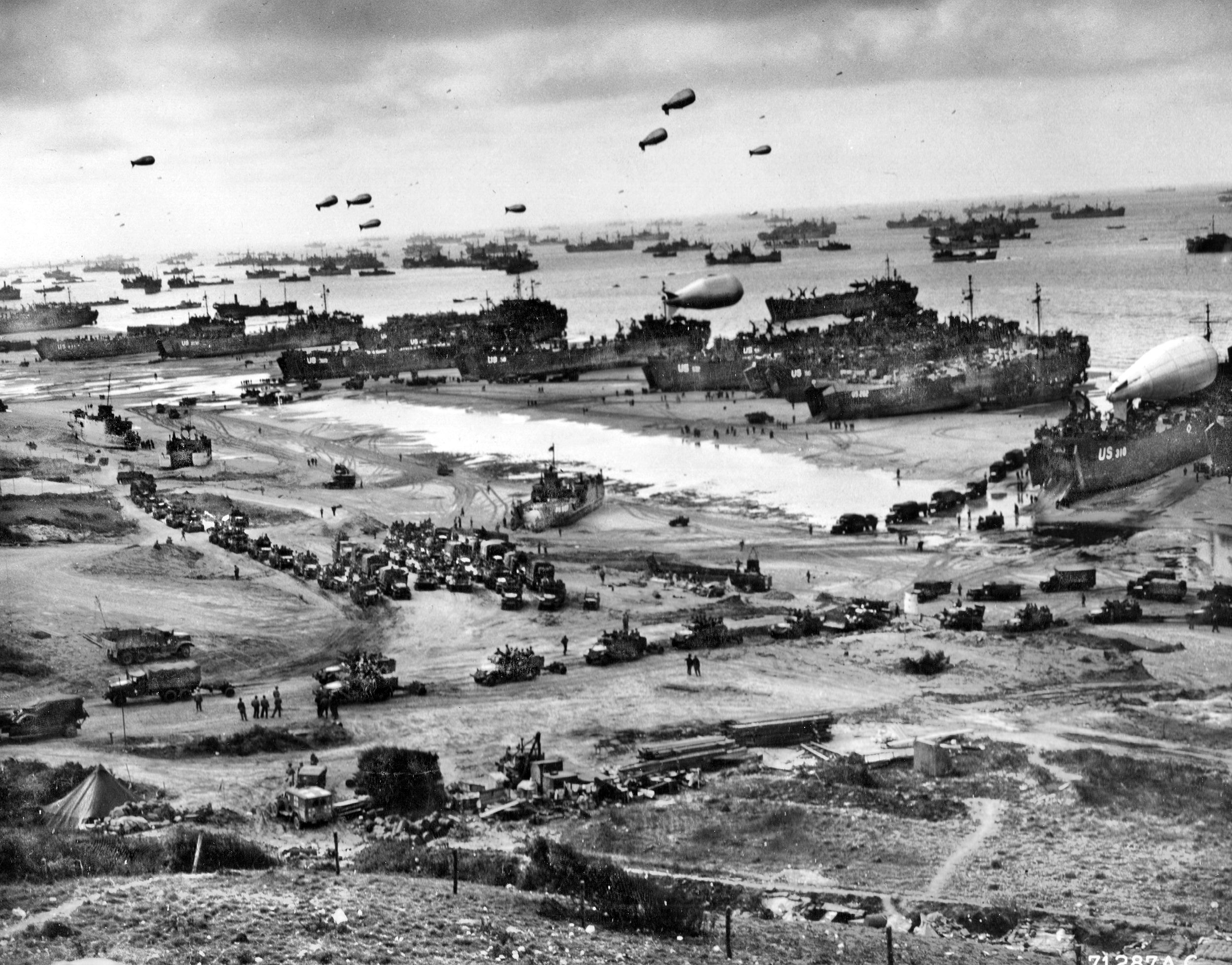 Barrage balloons float above landing craft and other vessels clustered at Omaha Beach shortly after the D-Day landings. The balloons, handled by the 320th Barrage Balloon Battalion, were intended to guard against attacks from low-flying enemy aircraft. At far right a barrage balloon rests on the deck of an LST (Landing Ship, Tank) prior to deployment.