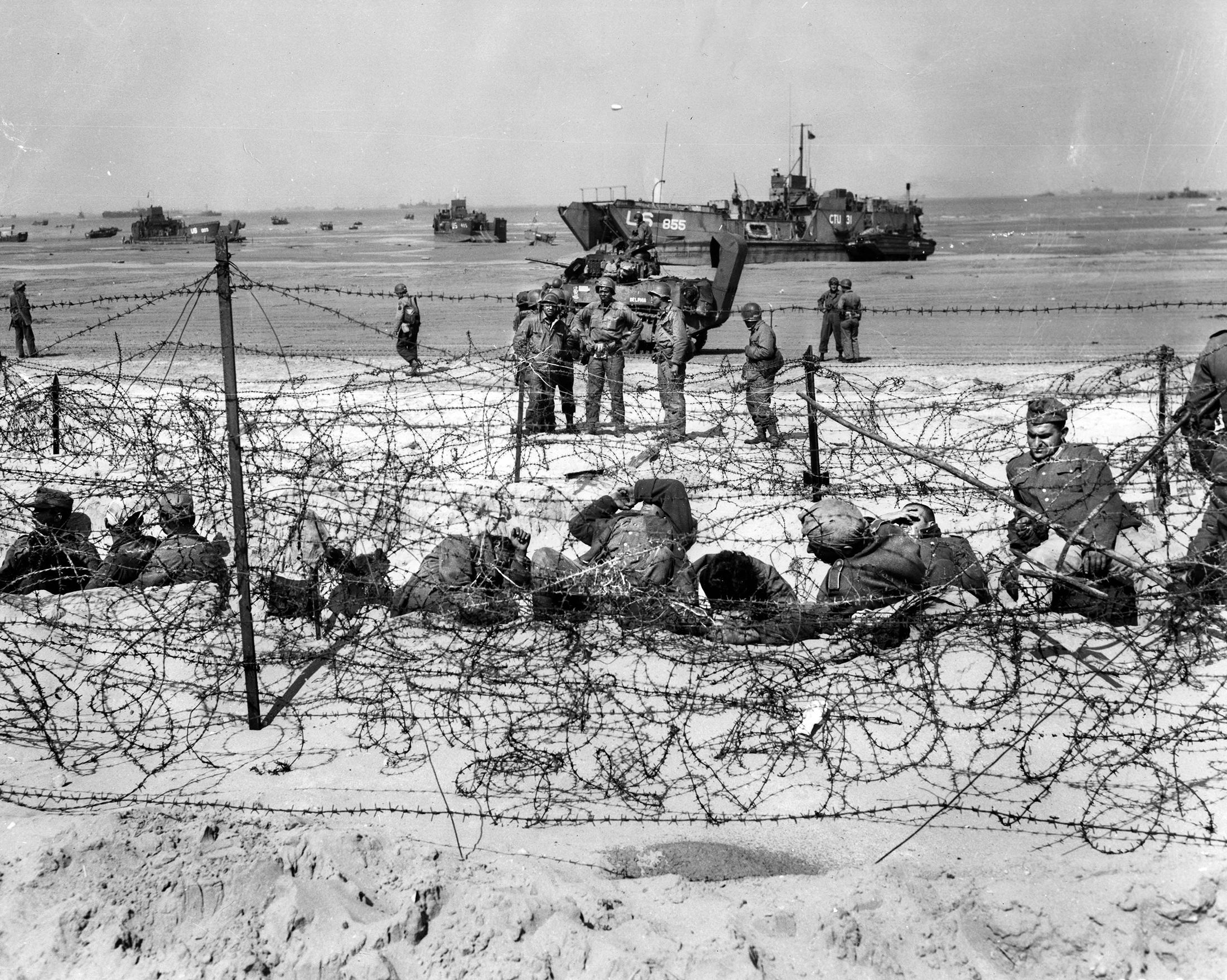 U.S. soldiers of the 320th Barrage Balloon Battalion, an African American unit, are seen standing in the background of this photo of German prisoners inside a barbed wire enclosure at Utah Beach on the afternoon of D-Day. 