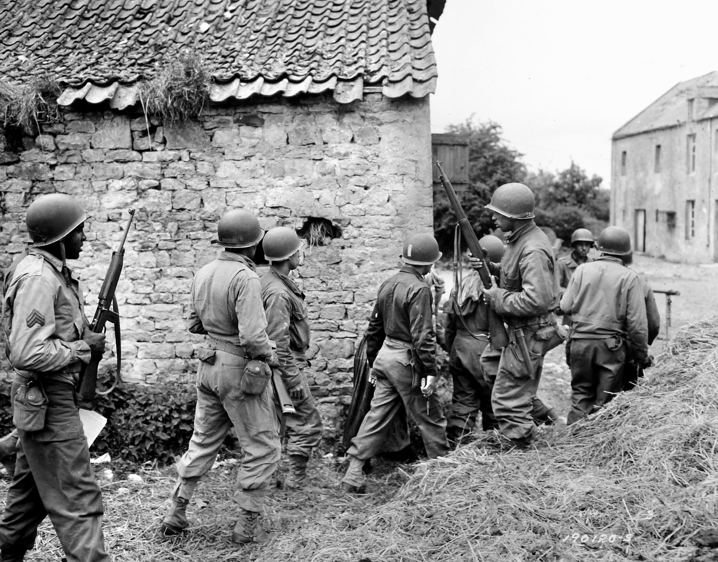 On June 10, 1944, D-Day+4, African American soldiers of the 320th Barrage Balloon Battalion search for German snipers in the French village of St. Laurent-sur-Mer. Visible at center with the recognition stripe on his helmet is a white platoon officer accompanying the soldiers.