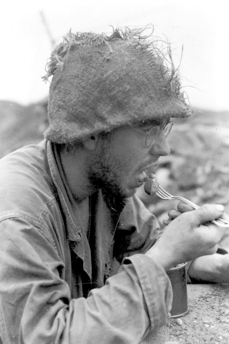 An infantryman of the 2nd Division with a sandbag helmet cover, enjoys a meal.