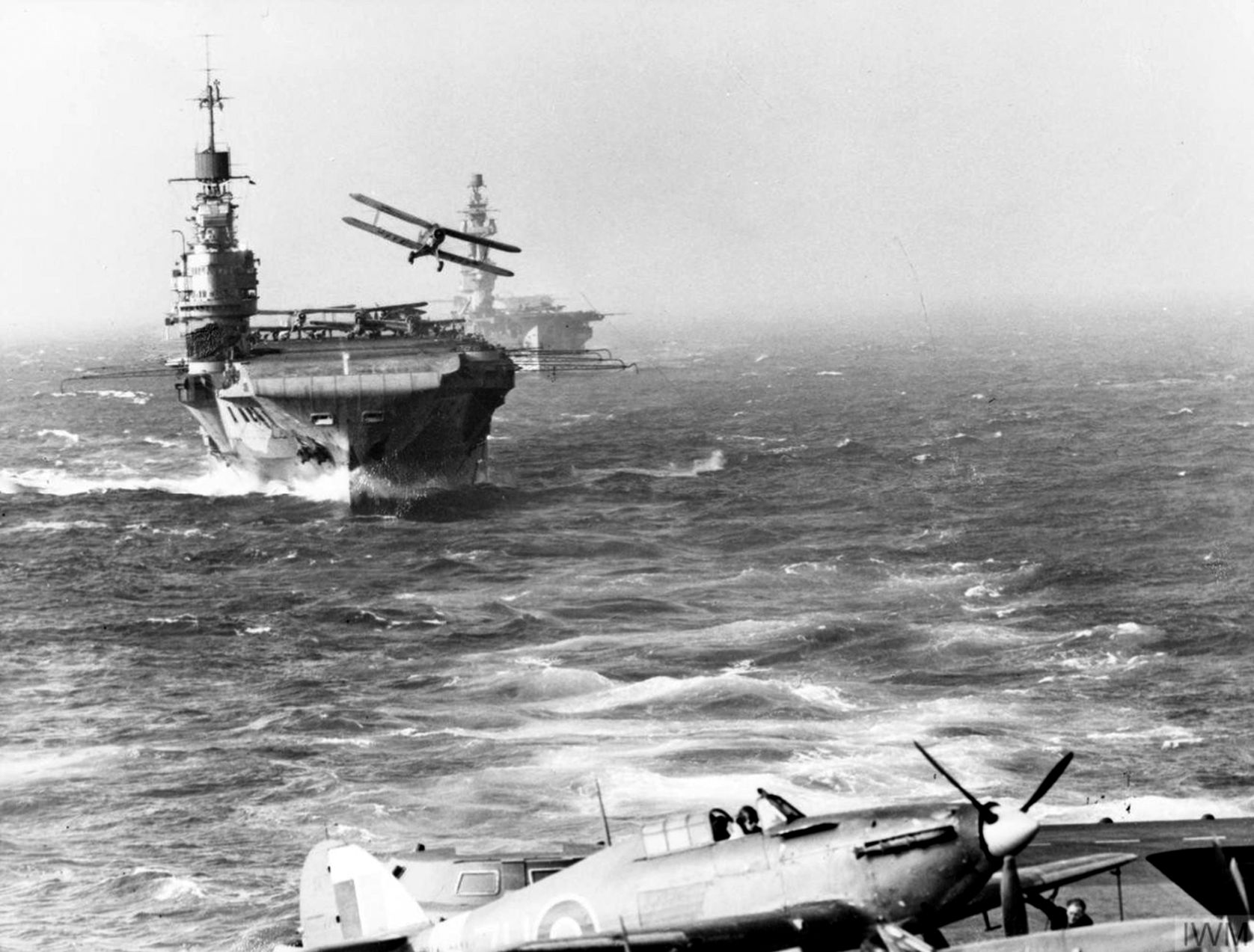 Taken from the aircraft carrier HMS Victorious, this photo shows a Hawker Hurricane fighter on the deck of Victorious while a Fairey Albacore biplane torpedo bomber takes off from the deck of the aircraft carrier HMS Indomitable, and the aircraft carrier HMS Eagle is shown in the background. This image was taken at sea in August 1942.