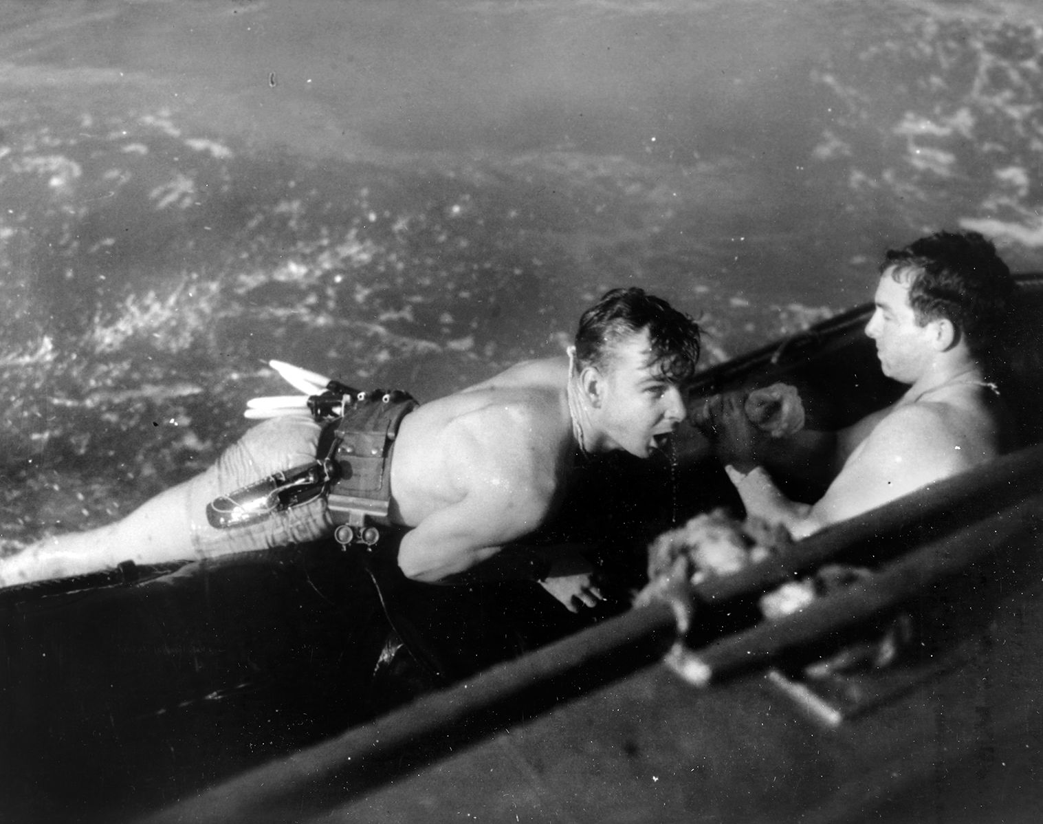 A member of an Underwater Demolition Team (UDT) is brought aboard a rubber raft that is towed by a powered watercraft. Note the life belt that holds the diver’s knife in its sheath and other basic tools.