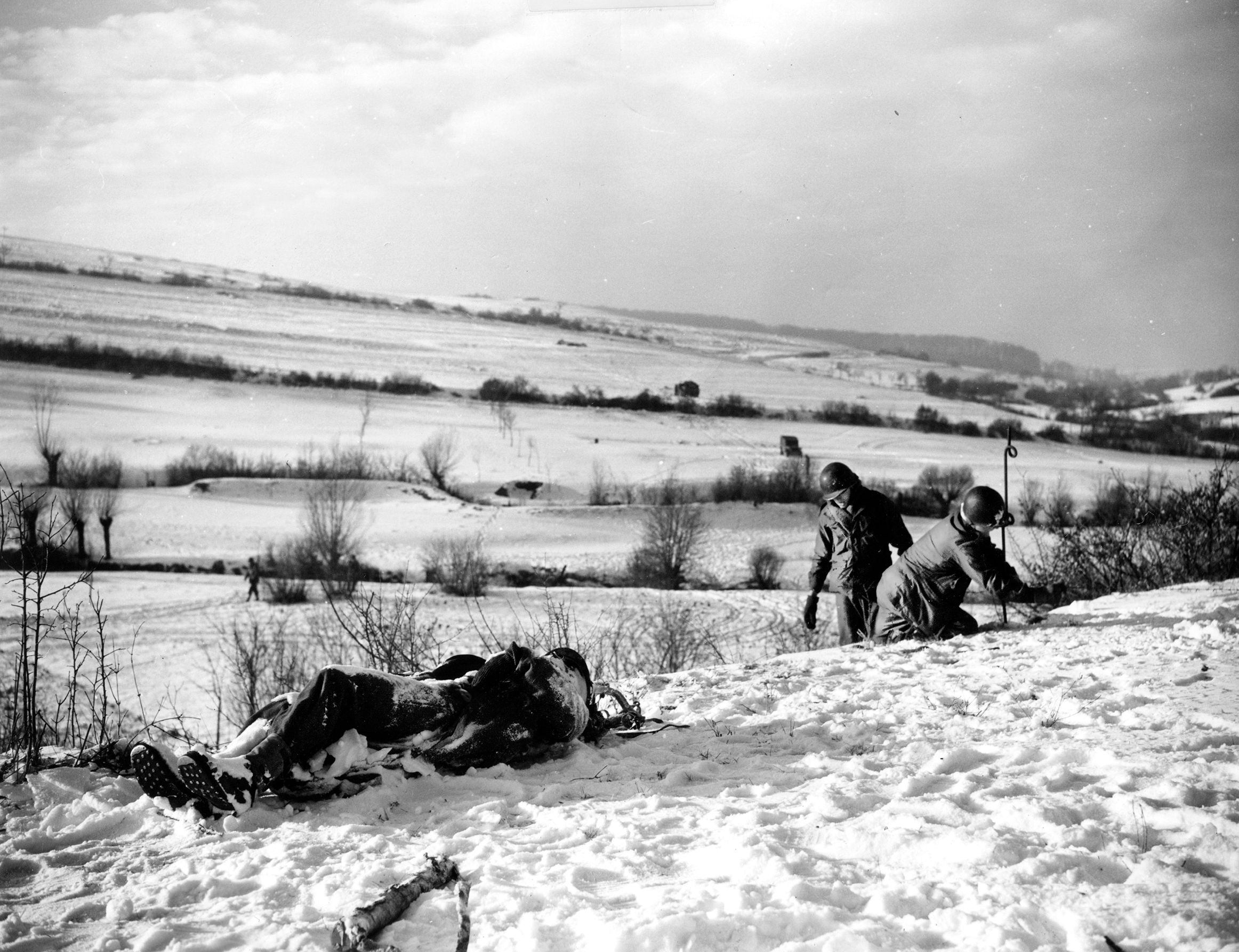 Engineers of the U.S. 100th Infantry Division string barbed wire to secure positions against enemy attack. They appear either undisturbed by the presence of a dead German's body lying nearby in the snow or oblivious to it. 