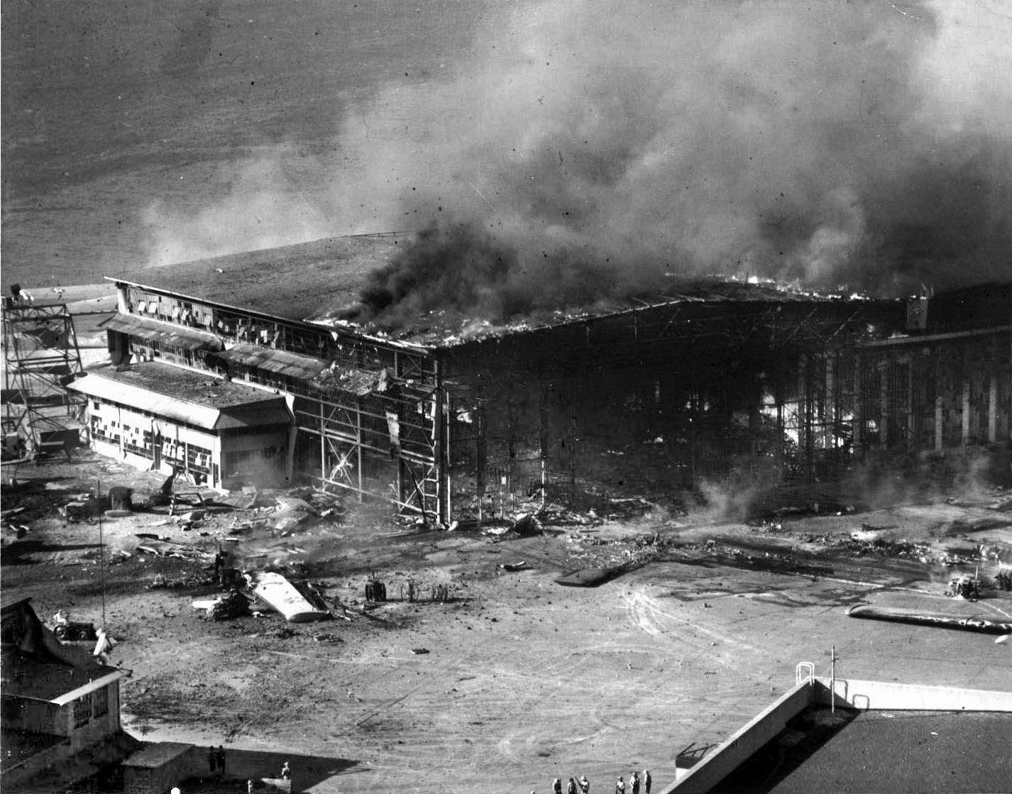 Debris from shattered Consolidated PBY Catalina flying boats destroyed on the ground litters the area at Ewa Marine Corps Air Station on December 7, 1941. After taking a direct hit from a Japanese bomb, a hangar is fully engulfed in flames as smoke billows skyward. 