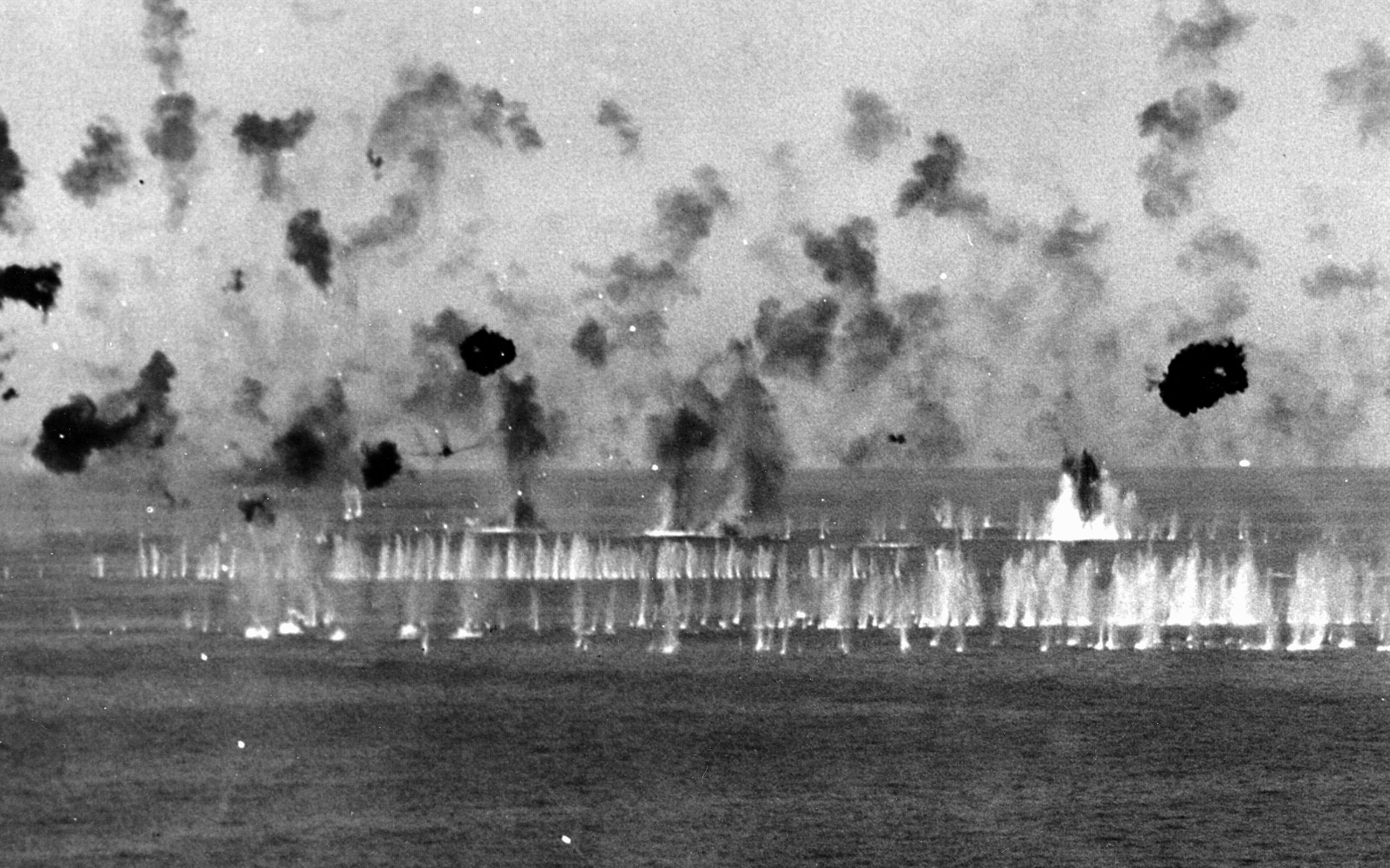 As antiaircraft shells churn the water around it, a Japanese “Kate” torpedo bomber (circled) presses home its attack.