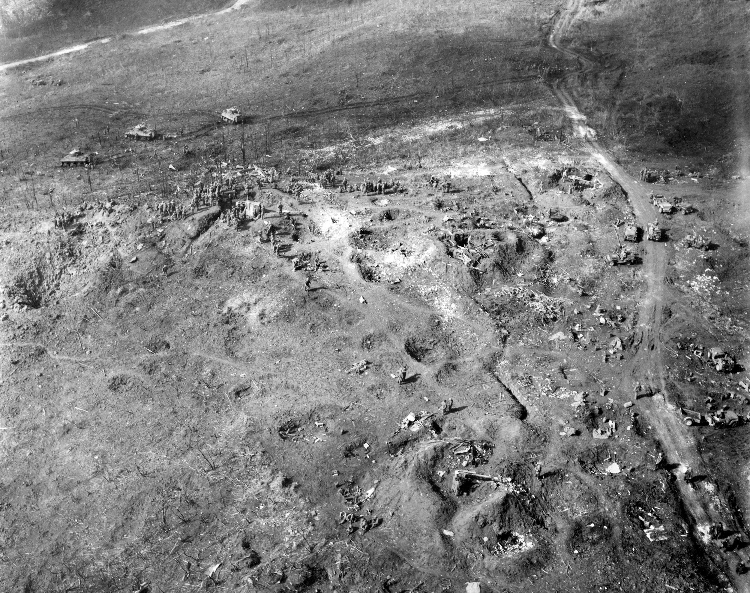 An aerial view of the area on the island of Saipan where theJapanese suicide attack (gyokusai) took place July 7, 1944. Many of the American survivors of the attack can be seen on the high ground near the top of the photo. Others are walking or standing around, with dead and possibly wounded men lying on the ground.