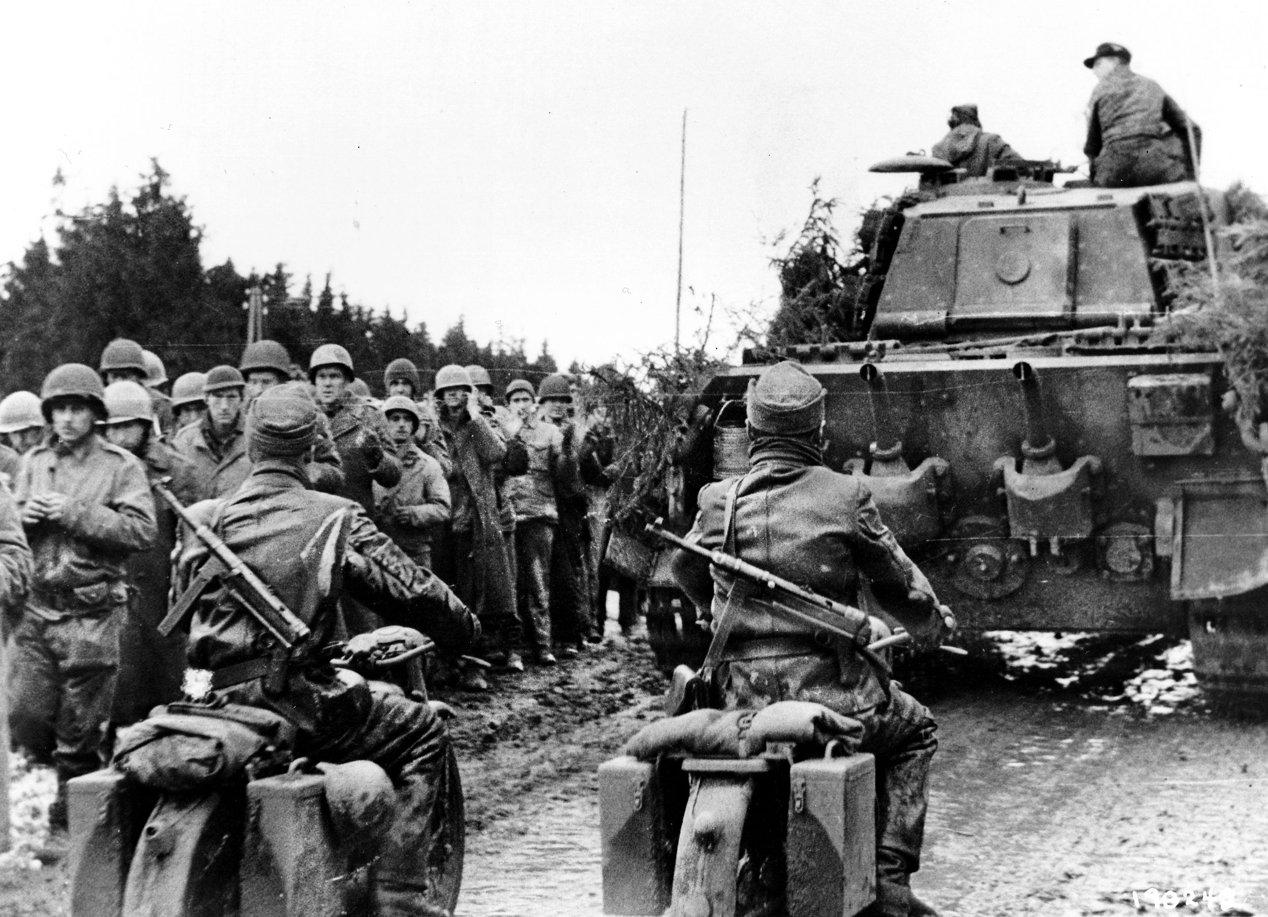 Two German motorcyclists armed with MP-40s follow a Königstiger tank as American POWs march along Rollbahn D towards Merlscheid, Germany.