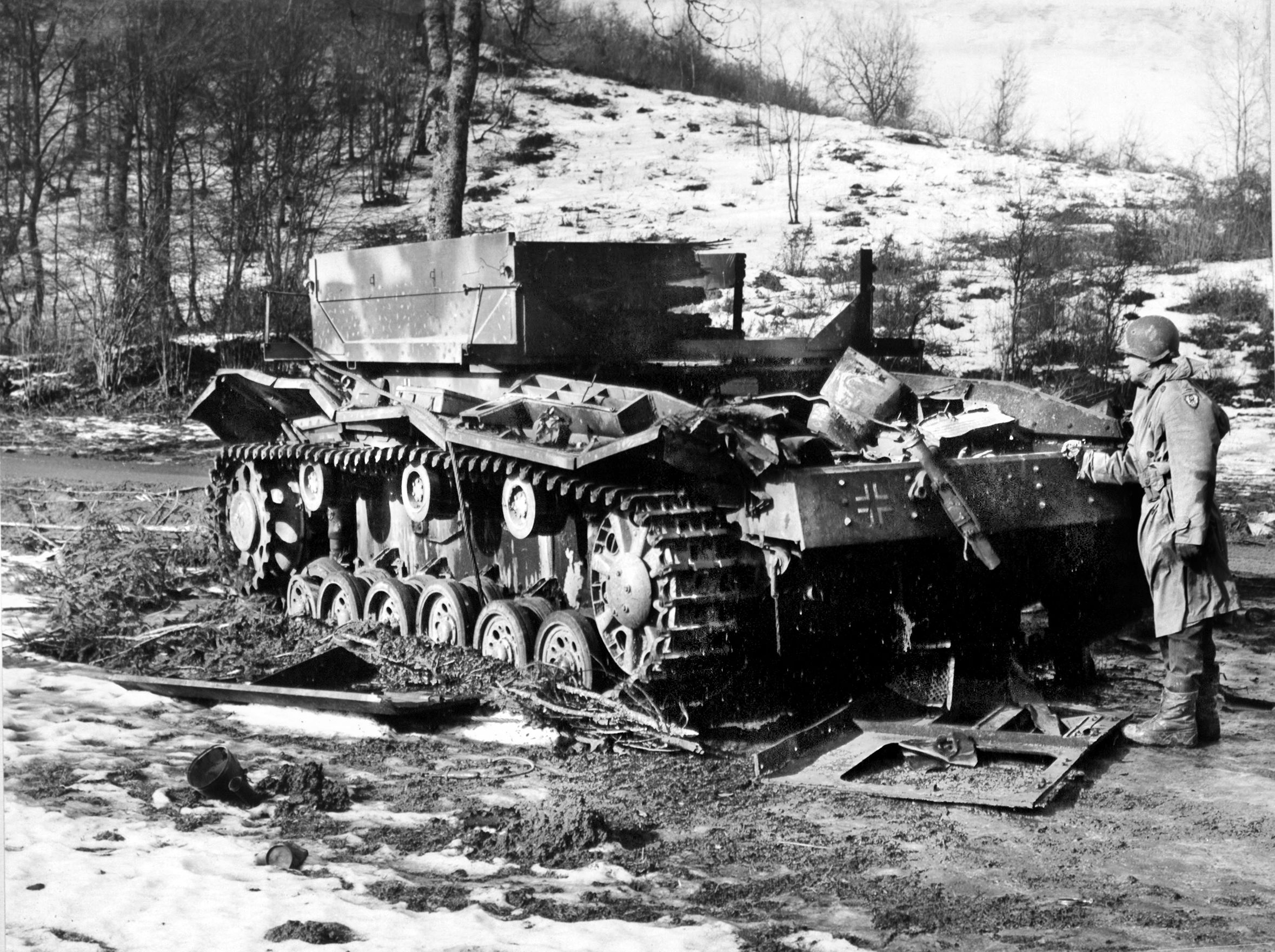 Weeks after the Ardennes forest fell silent, an U.S. soldier examines the charred remains of a German self-propelled gun on the outskirts of Hosingen, Luxembourg. The wreckage serves as a grim monument to the stubborn defense mounted by the 110th Infantry Regiment (28th ID), which turned this small crossroads town into a costly bottleneck for the German advance.
