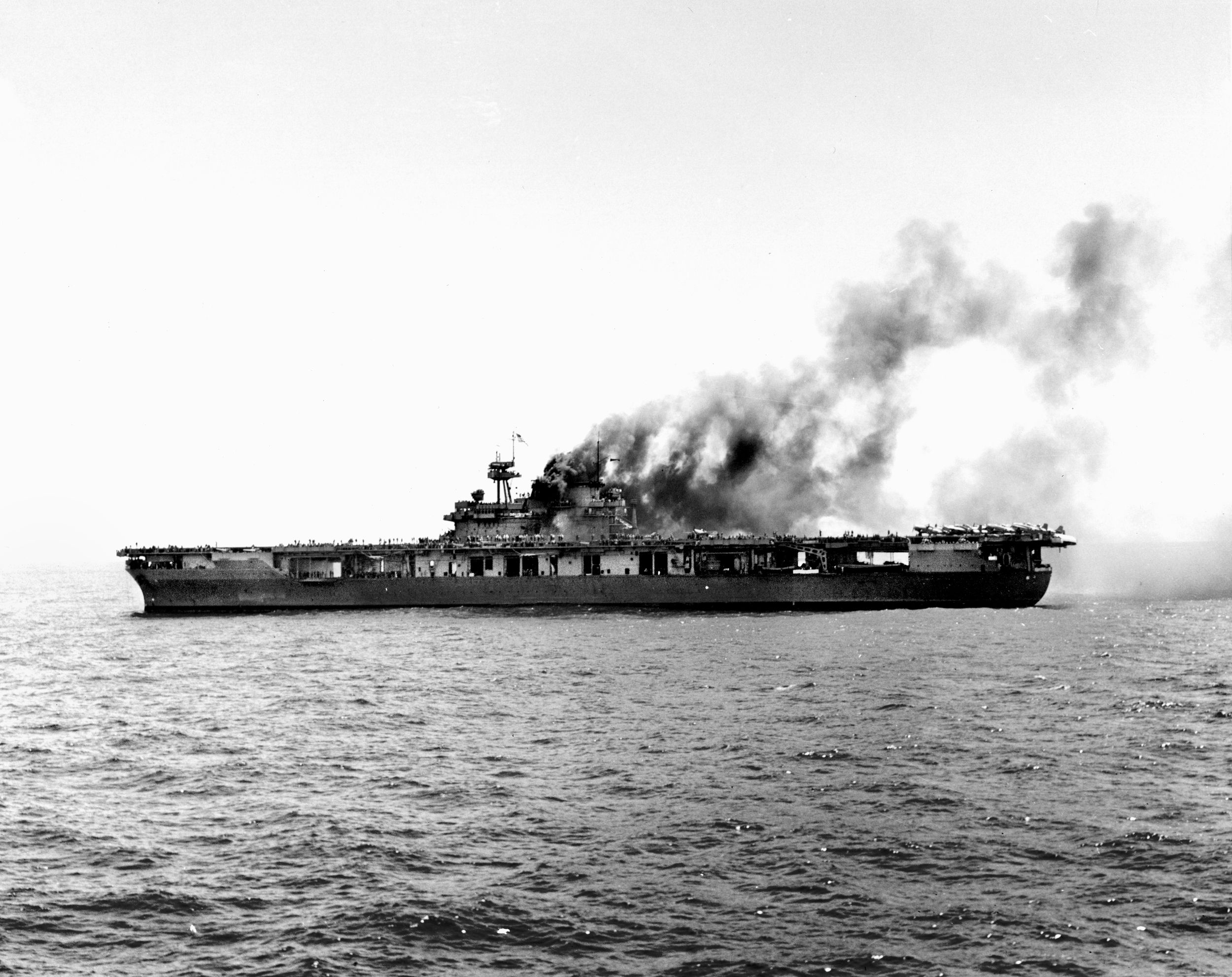 USS Yorktown sits dead in the water about an hour after the initial Japanese noon bomb attack at Midway on June 4, 1942. Though fires still smolder in her uptakes, immediate repairs are already well advanced; F4F-4 fighters have been respotted aft for takeoff, and two SBD-3 scout bombers are visible in the hangar bay below. This spectacular damage-control effort got the carrier back underway at 20 knots, just before a second Japanese strike wave targeted her with two fatal aerial torpedoes.