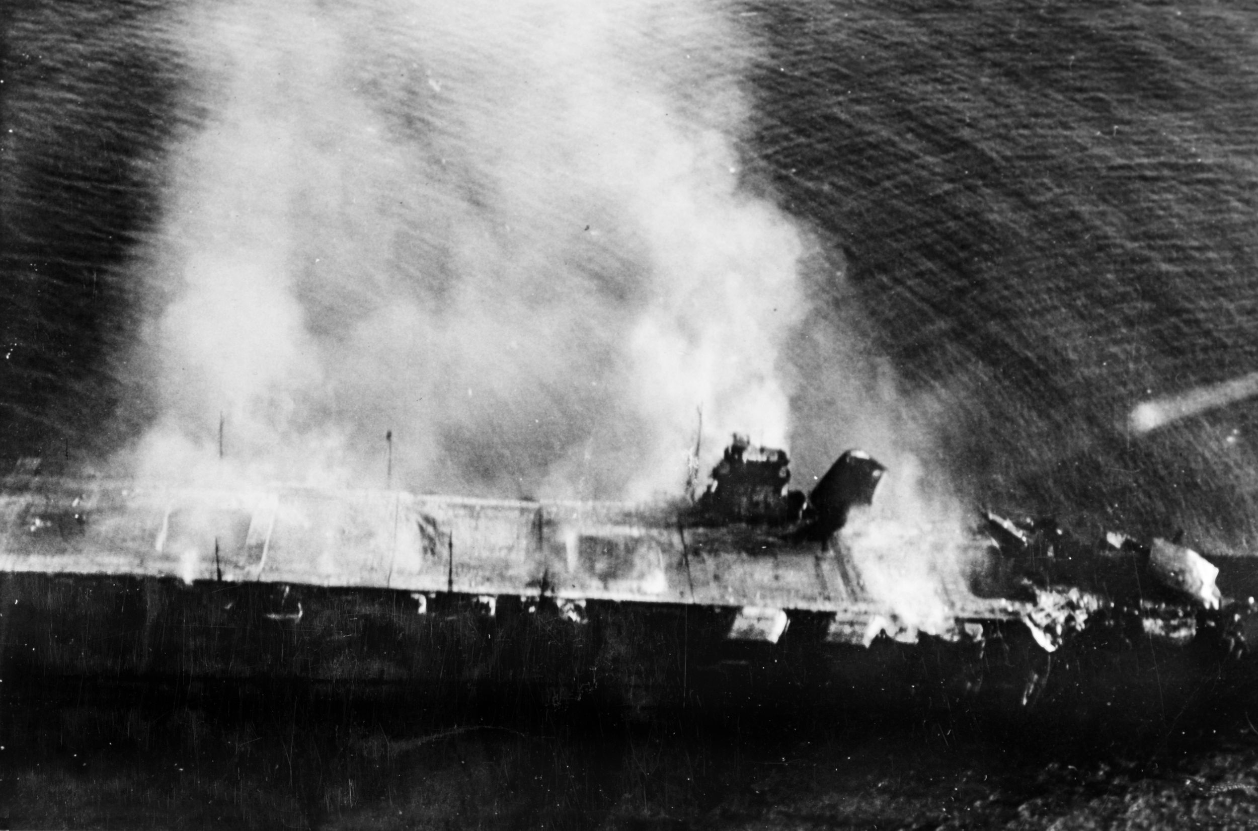 A plane from the carrier Hosho captured this image of the burning wreck of the Japanese aircraft carrier Hiryu adrift in the Pacific just after sunrise on June 5, 1942. U.S. Navy dive-bombers have done their work—the flight deck on the right has completely collapsed, and a section of the forward elevator stands vertically upright directly in front of the island, thrown into the air by a devastating internal hangar explosion.