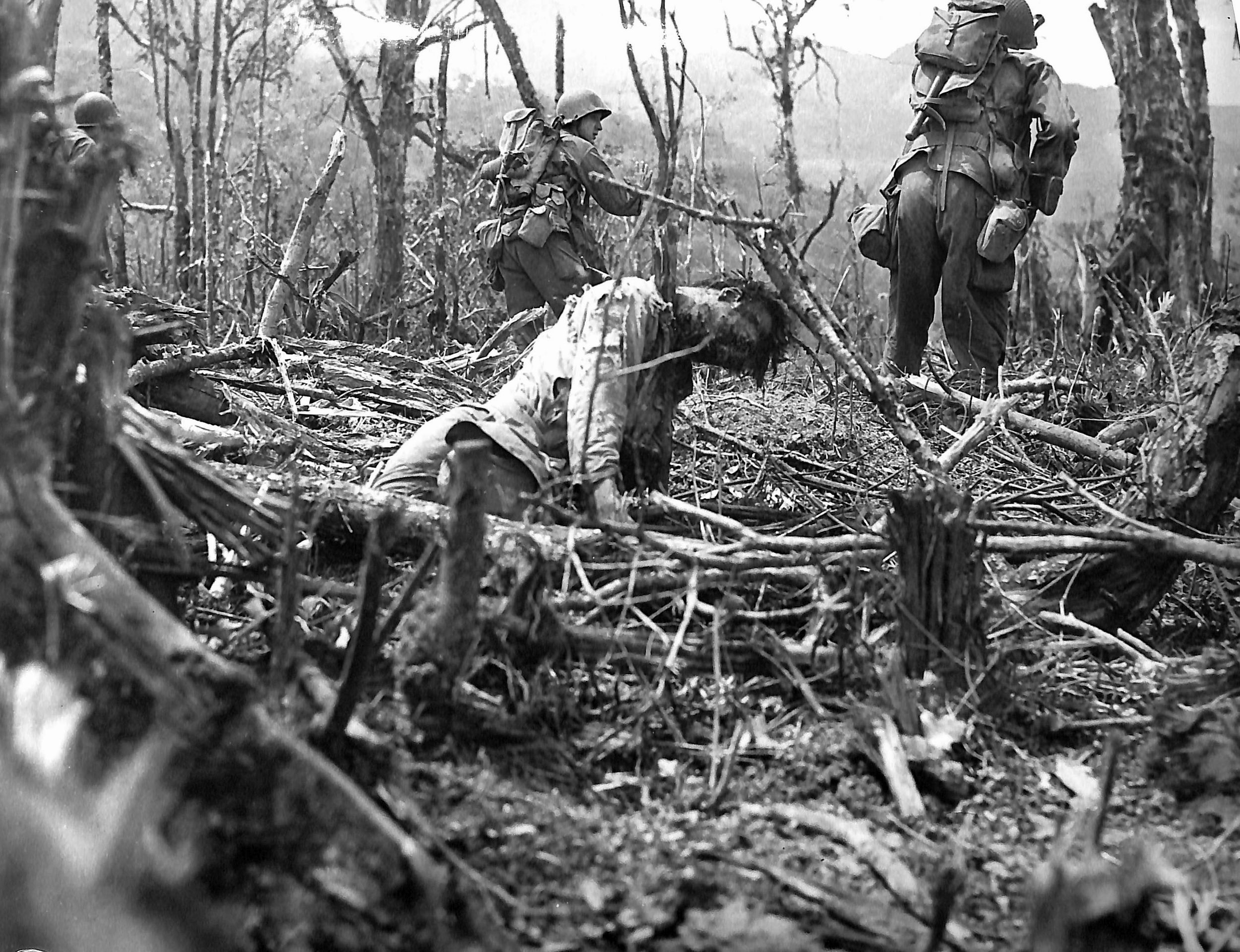 Battle-weary infantrymen of the 25th Infantry Division (“Tropic Lightning”) bypass a fallen Japanese defender as they scale a scarred, shell-torn ridge near Balete Pass in the spring of 1945. Dubbed “Bloody Pass” by the troops who fought there, the critical mountain gateway was defended by thousands of fanatical Japanese soldiers who contested every ridge, bunker, and reverse-slope position to the death.
