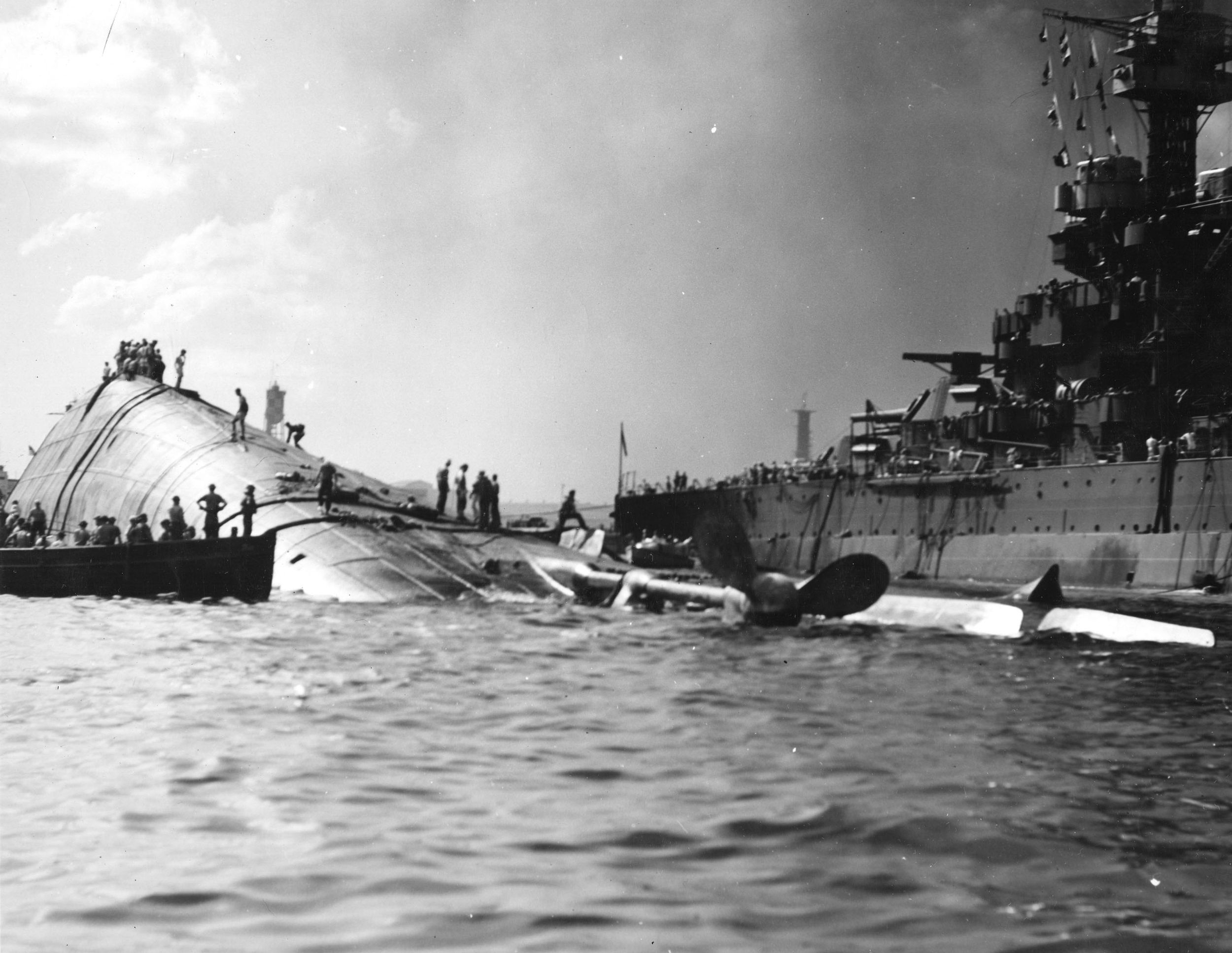 The capsized hull of the battleship USS Oklahoma lies in the shallow water of Pearl Harbor. The Oklahoma was hit by several Japanese torpedoes, causing the ship to capsize and trapping a number of sailors below decks. Feverish efforts to rescue the trapped men were only partially successful.