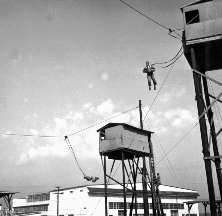 A Canadian airborne candidate exits the most dreaded apparatus in paratrooper training—the 35-foot mock tower—during pre-invasion training. Used to build muscle memory for a rapid exit and proper body position, this towering structure was a psychological hurdle that broke many volunteers before they ever went up in a real plane.