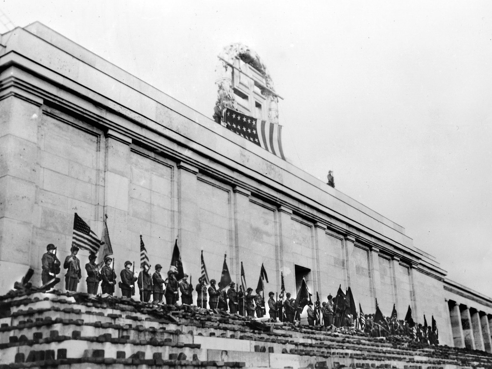 An American flag covers the Nazi swastika atop the facade of Nuremberg’s Kongresshalle—a massive, unfinished structure designed to resemble the Roman Colosseum. Following the capture of the “City of the Rallies” on April 20, 1945, soldiers of the 45th Infantry Division assembled in the shadow of the building, whose die-hard defenders had to be cleared floor-by-floor.