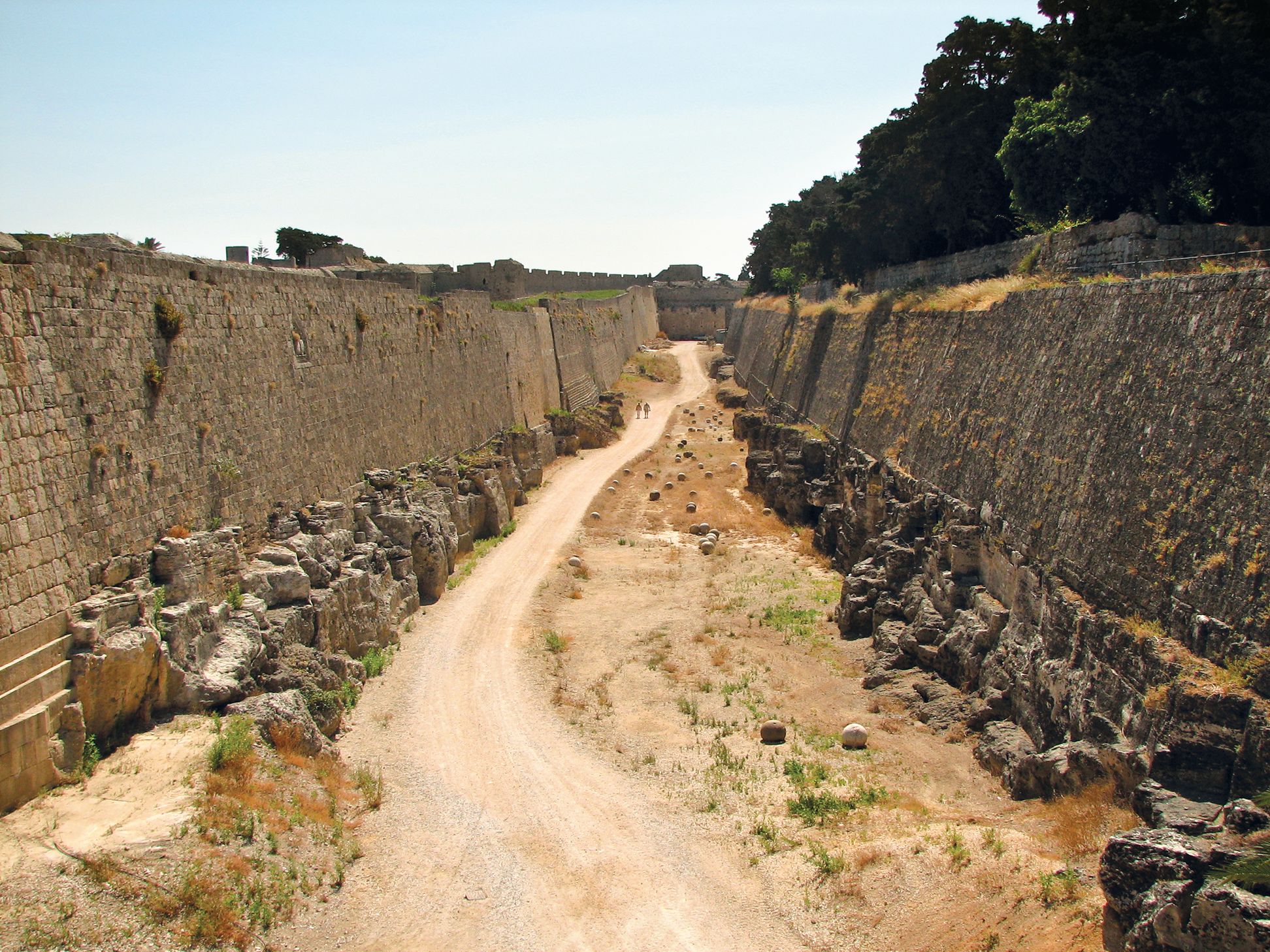 The English Post section of Rhodes’ medieval city walls as seen today. This was the scene of heavy bombardment and intense combat during the Ottoman siege of the Knights Hospitaller in 1522. The crenelated city walls can be seen in the background behind the tenaille—a defensive-work, in front of the main walls of a fortress—on the left. Across the ditch is the counterscarp, another obstacle for attackers trying to reach the main wall. The stone cannonballs to the right of the road are from the time of the siege.