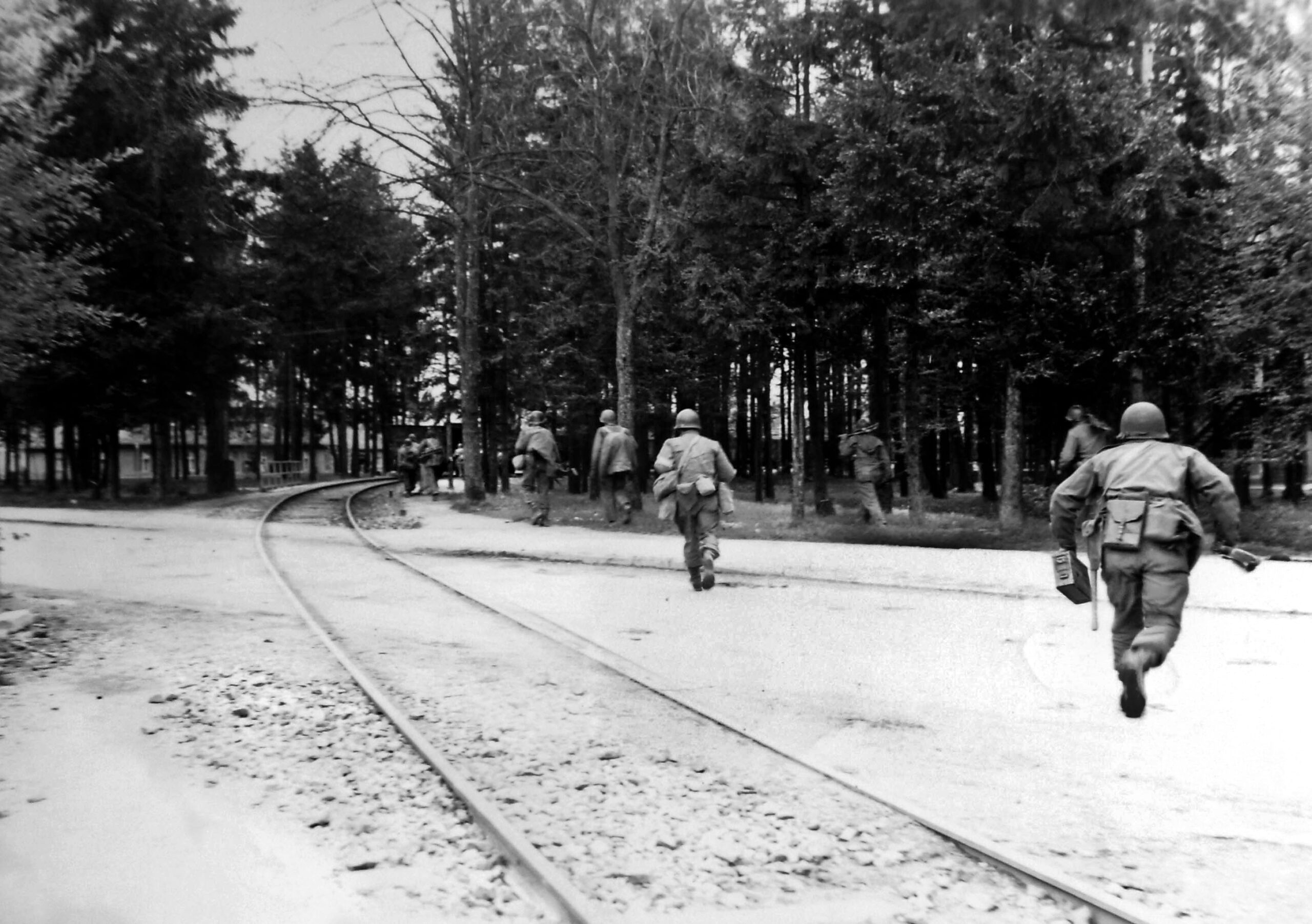 American troops, likely elements of the 45th “Thunderbird” Infantry Division, advance under sporadic fire from SS guards as they approach the Dachau concentration camp complex near Munich on April 29, 1945. Barely visible through the trees is part of the infamous “death train” on which thousands died from starvation, exhaustion, or execution. In an effort to evacuate prisoners as the Allies approached Buchenwald, the SS crammed roughly 4,000 to 4,500 inmates into open and closed rail cars for a catastrophic 21-day journey to Dachau with little food or water.