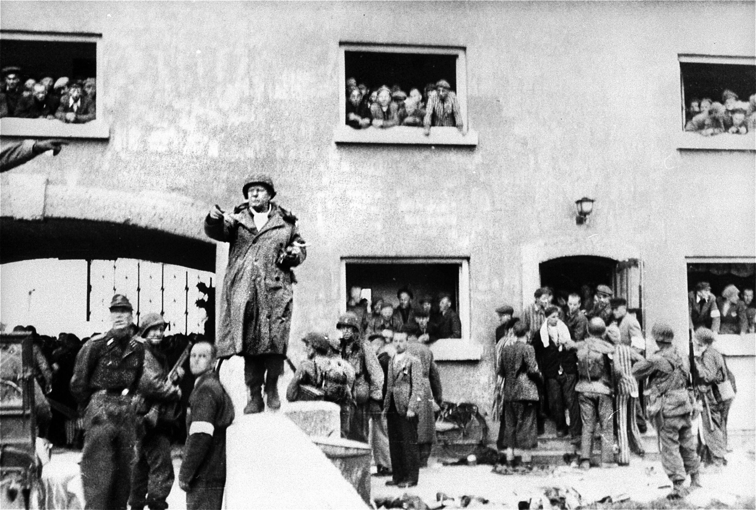 Brigadier General Henning Linden, assistant division commander of the 42nd “Rainbow” Infantry Division, stands outside the Jourhaus (gatehouse) at the main entrance to Dachau on April 29, 1945. Standing below him is Dr. Victor Maurer of the International Committee of the Red Cross (wearing white armband), alongside SS-Obersturmführer Heinrich Wicker (far left), the German officer who formally surrendered the concentration camp to U.S. forces. In the background, liberated inmates watch the historic scene unfold from the gatehouse windows. This portal, through which inmates entered the prison, featured a wrought-iron gate bearing the cruel lie: ARBEIT MACHT FREI (“Work sets you free”). The cynical slogan offered false hope, as the primary reality of Dachau was death by starvation, disease, forced labor, or execution.