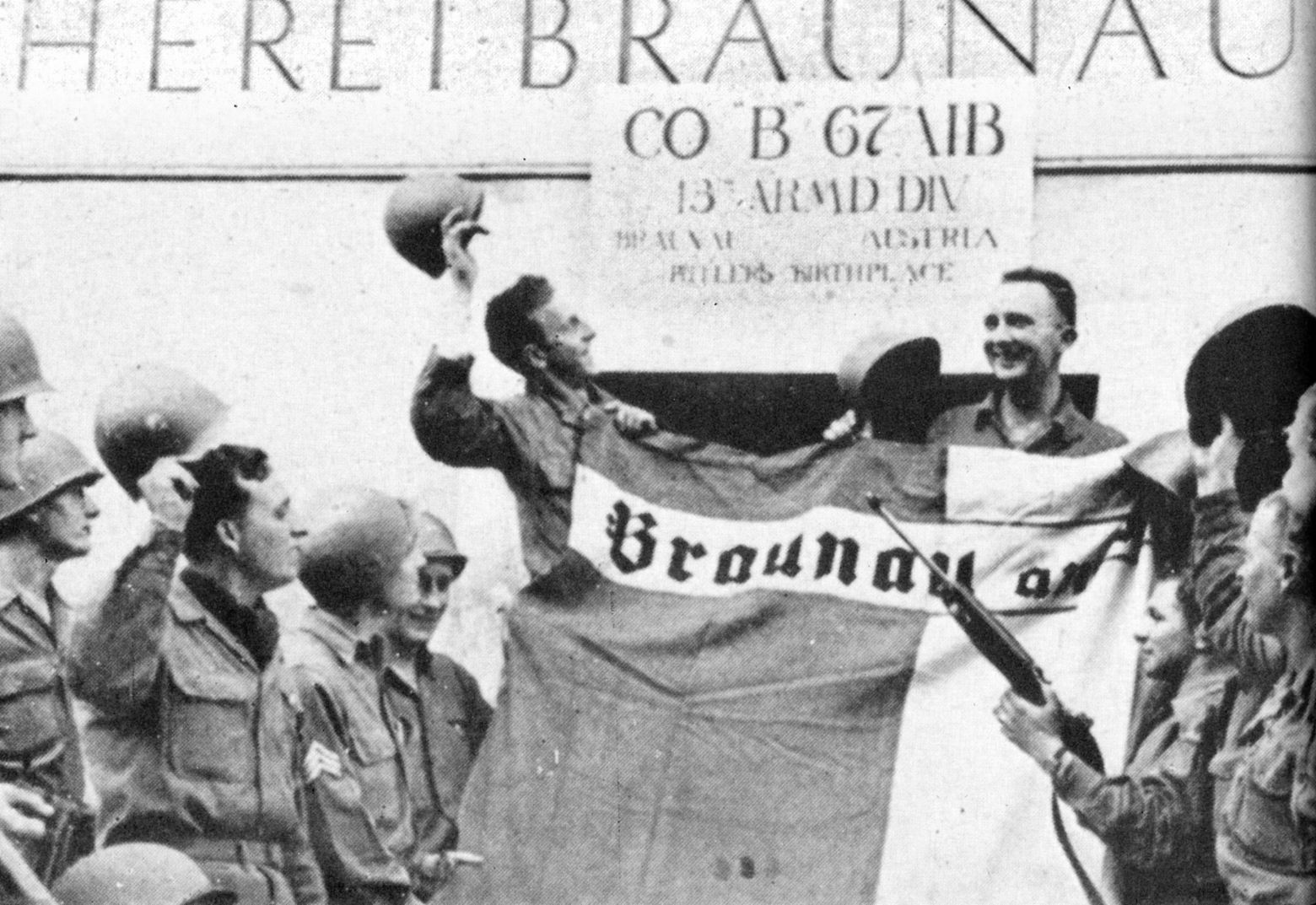 Soldiers from Company B, 67th Armored Infantry Battalion, tip their helmets toward Adolf Hitler’s birthplace in Braunau am Inn, Austria, while displaying a liberated city flag. The officer visible at the top left is likely the battalion commander, Lt. Col. Ridgway P. Smith.