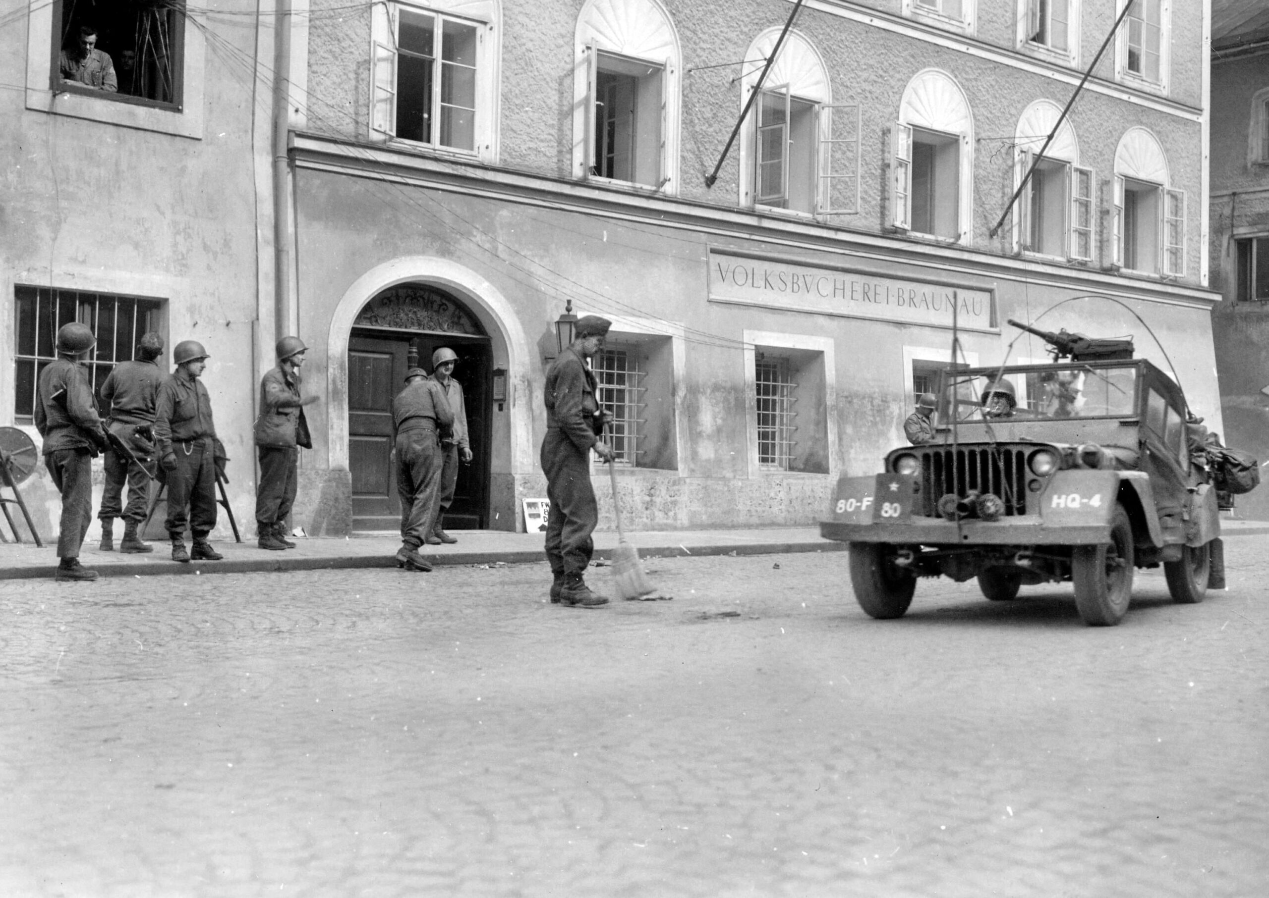Major General Horace McBride, commander of the 80th Infantry “Blue Ridge” Division, rolls past Adolf Hitler’s birthplace in Braunau am Inn, Austria, in an armed jeep on May 5, 1945, while a former German soldier sweeps away debris. Following their entry into the town, McBride’s division—part of Gen. George S. Patton’s Third U.S. Army—pushed rapidly southeast during the final 72 hours of the war in Europe, liberating concentration camps, accepting mass enemy surrenders, and linking up with Soviet forces before the official German surrender on May 8.