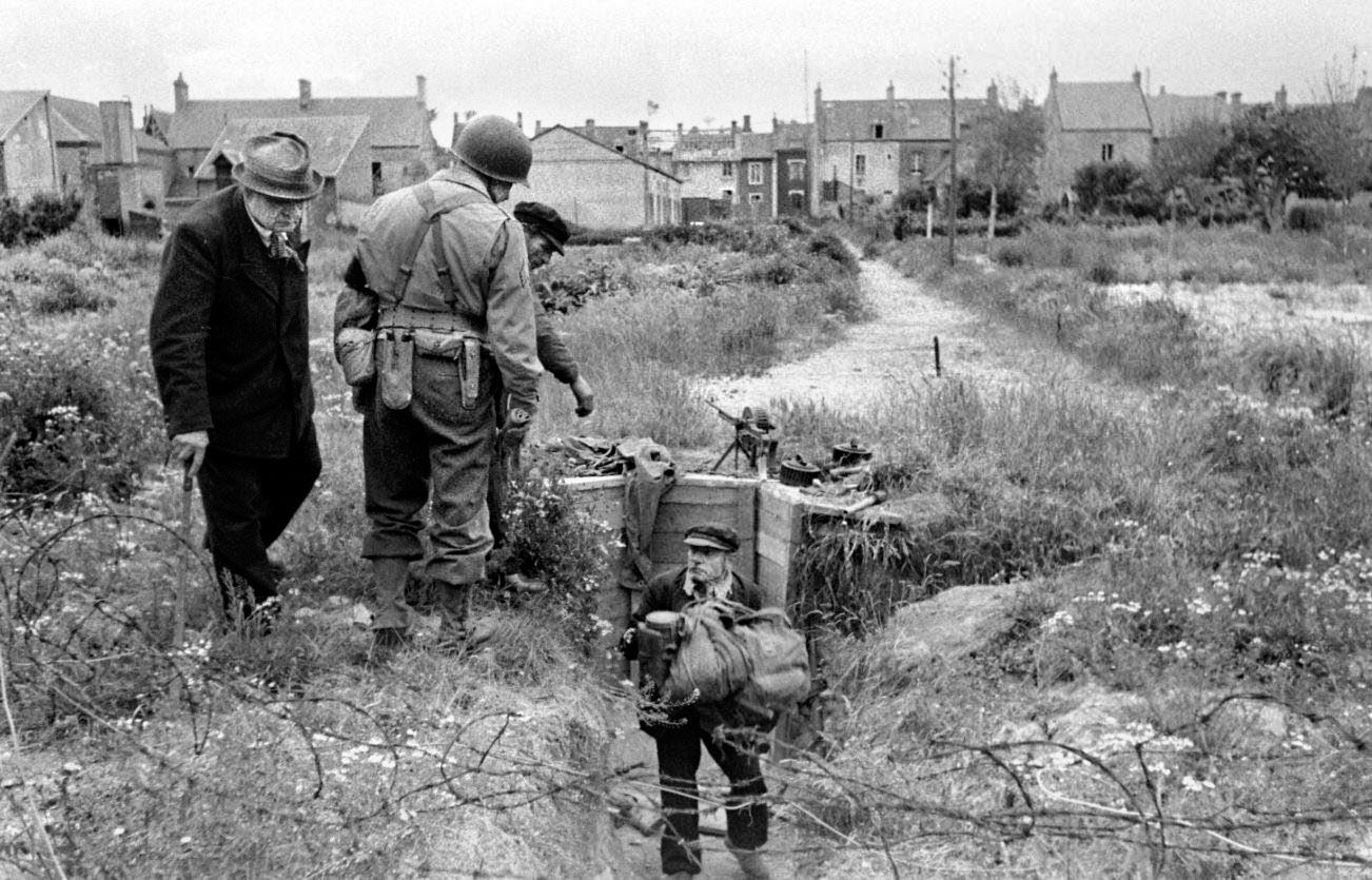 Counterintelligence Corps Sergeant Roland des Autels (back to camera) and Grandcamp town councilor Pierre Ferrari (left) watch as abandoned German equipment is cleared from a bunker near Maisy. In the background sits a French Reibel MAC 31 machine gun with its distinctive “camembert” drum magazine.