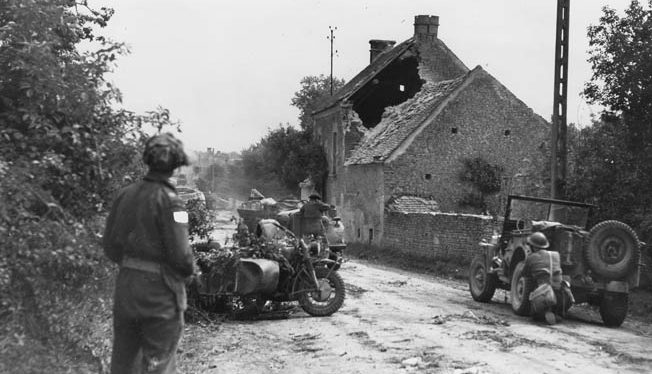 A Canadian patrol stops to inspect a German staff car and motorcycle in a village “somewhere in Normandy.” 